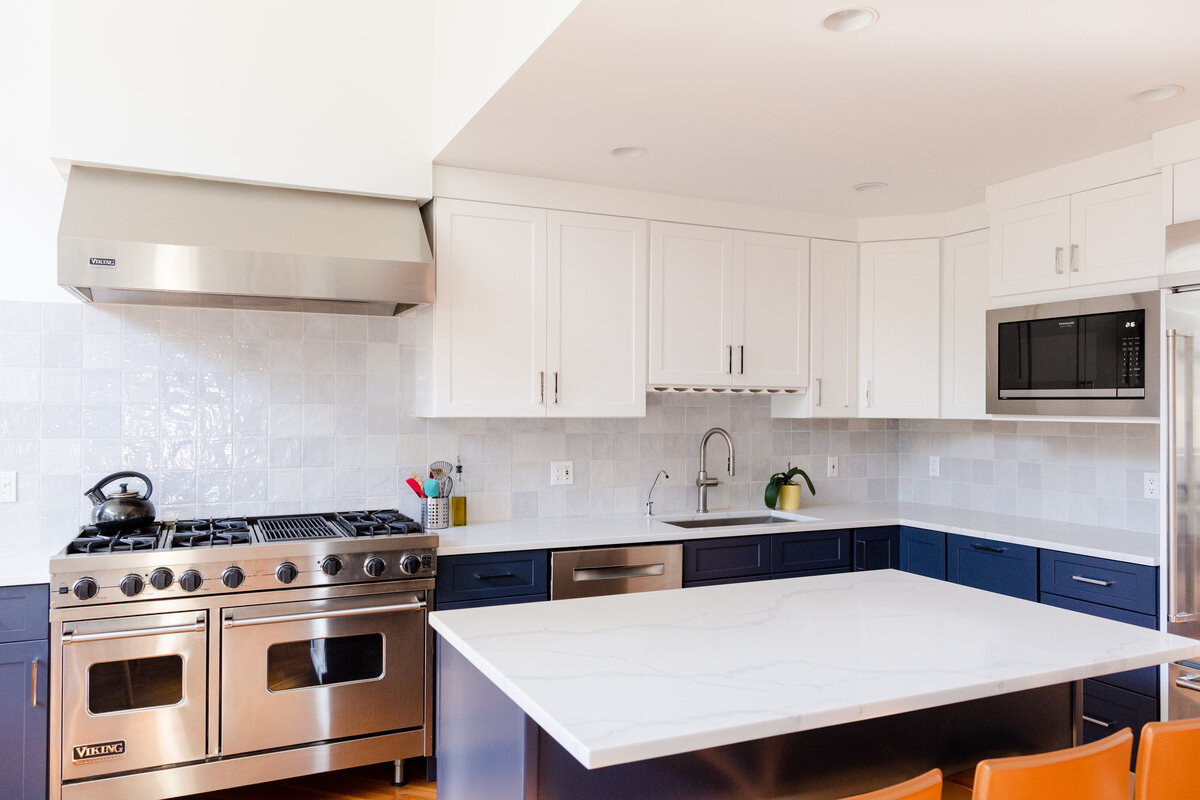 The kitchen island features two-tone blue and white cabinetry with steel appliances. Navy blue base cabinets are paired with frost white upper cabinets, and the walls are accented with blue ceramic zellige tile.