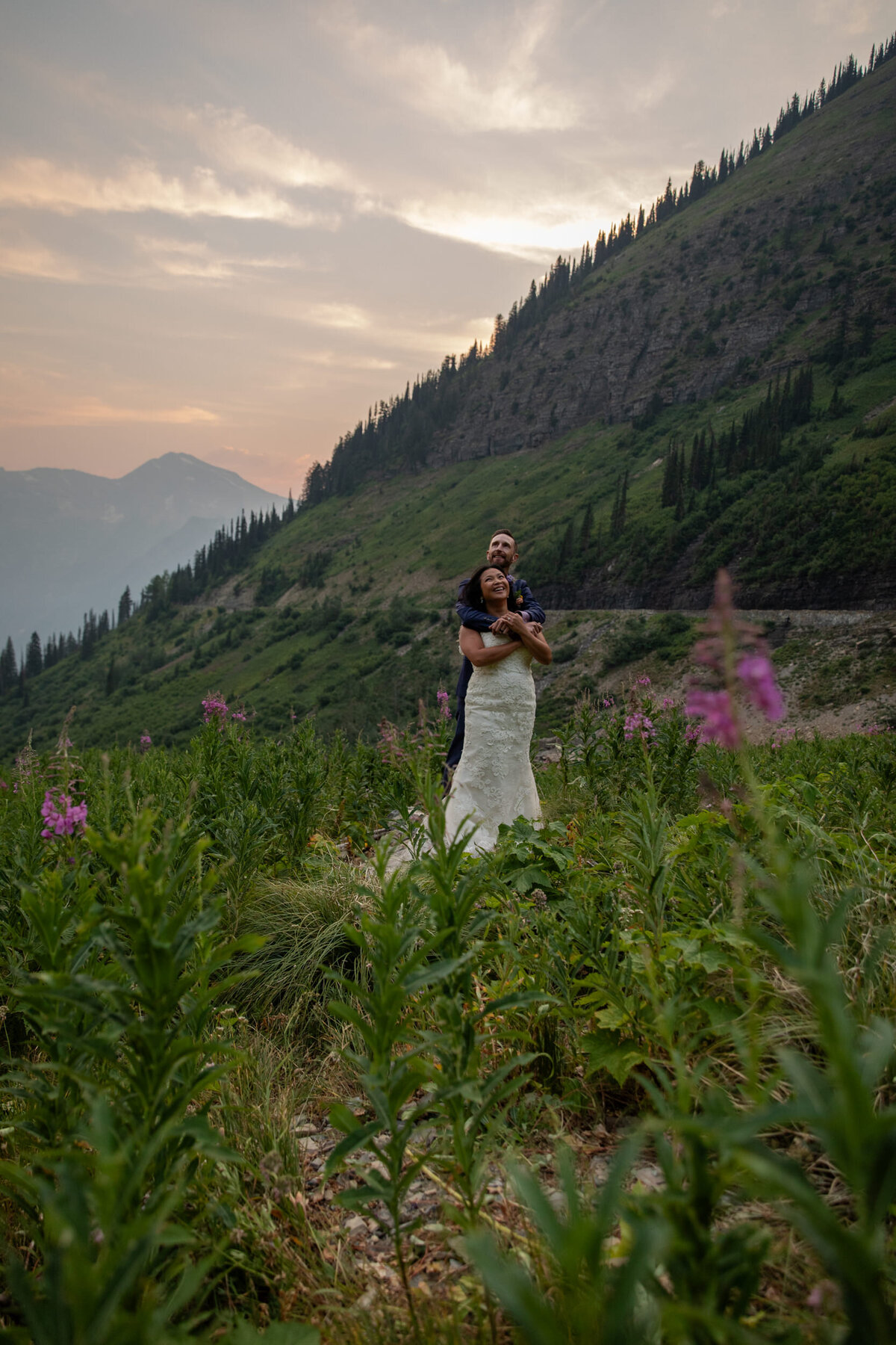 A bride stands in front of her groom as they laugh in a field of flowers.
