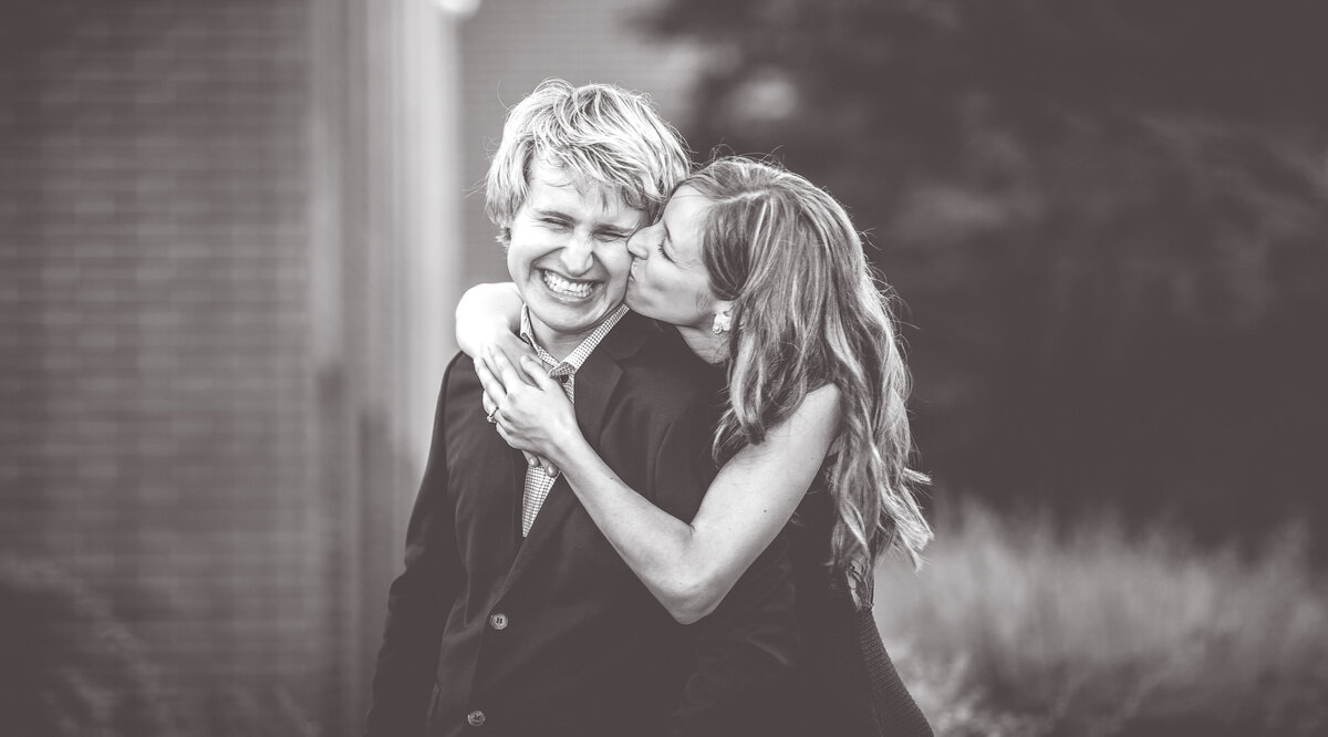 A cute photo of a bride kissing her groom while he is laughing during an outdoor engagement session.