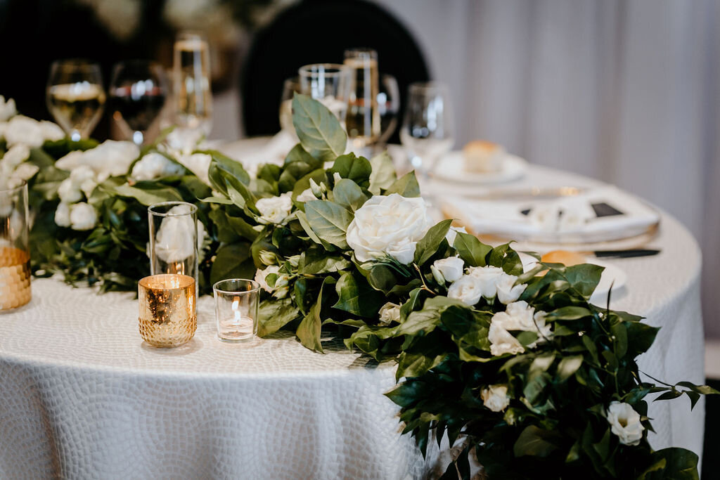 Elegant wedding table setup at the Palm Event Center in Pleasanton, CA, featuring a spring theme with a color scheme of gold, black, and white. The table is adorned with a floral runner of white blooms draping along crisp white linen, complemented by gold candleholders.