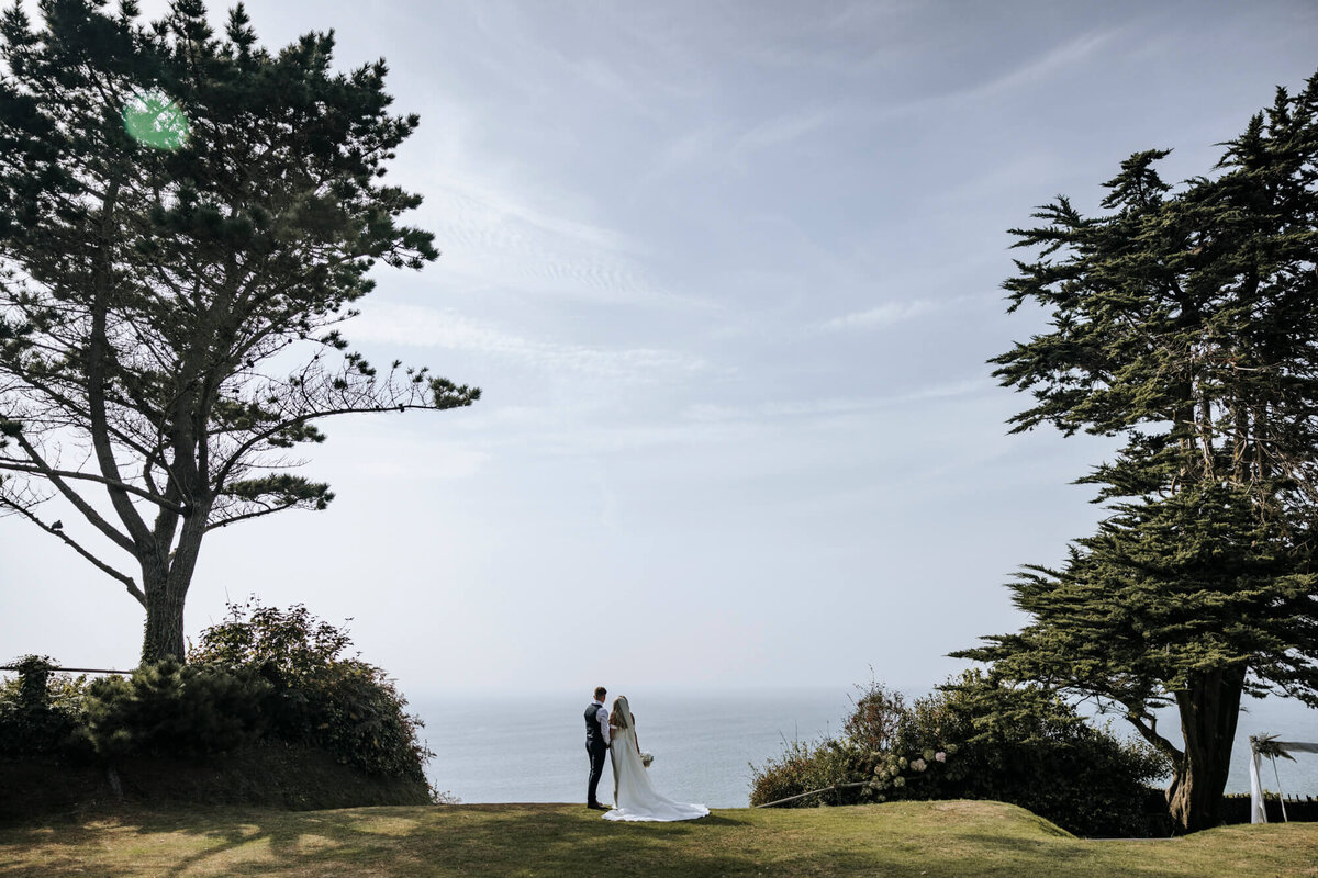 A bride & groom standing on top of a lush green hillside