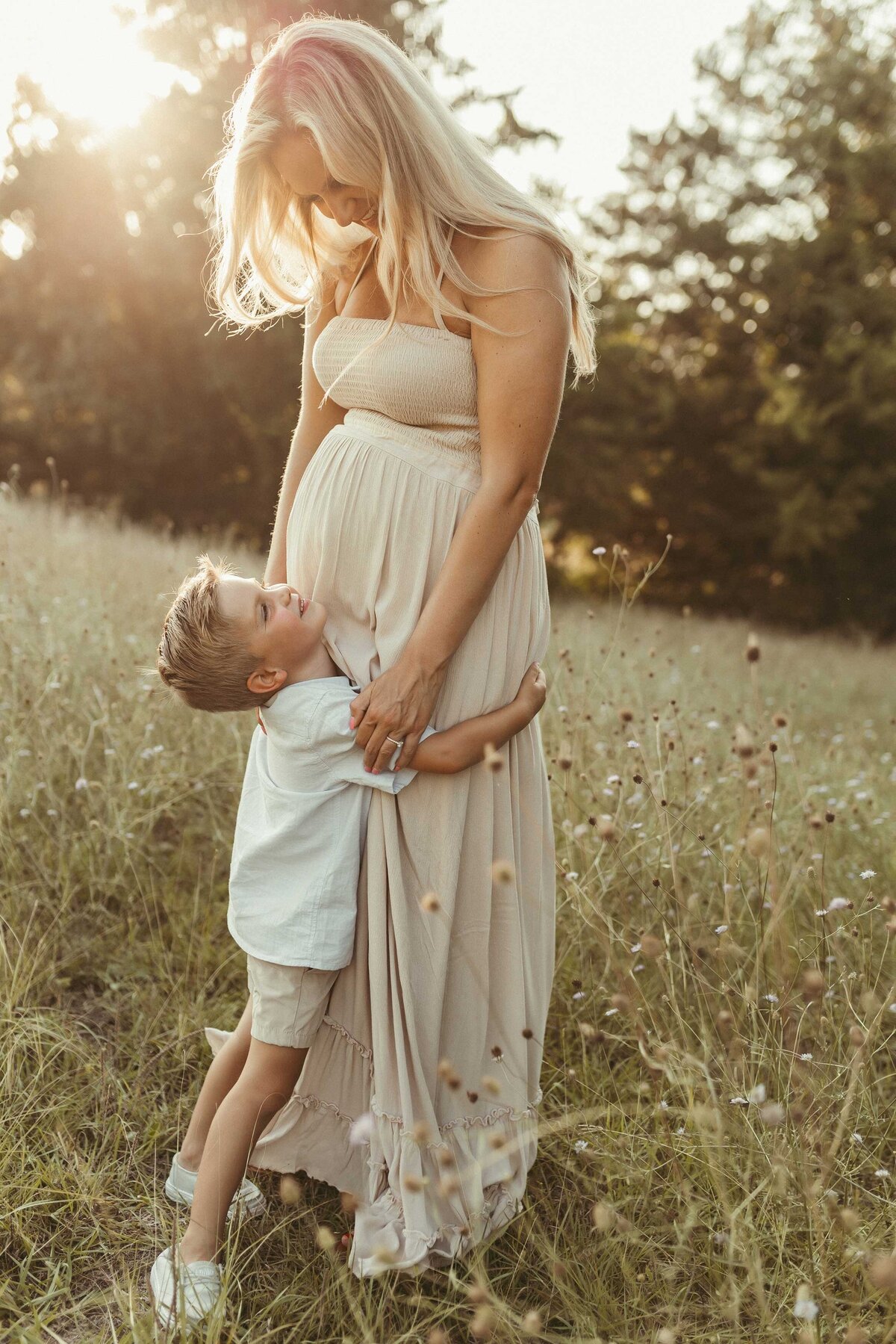 Blonde pregnant woman standing in a flower field with her young son hugging her hips