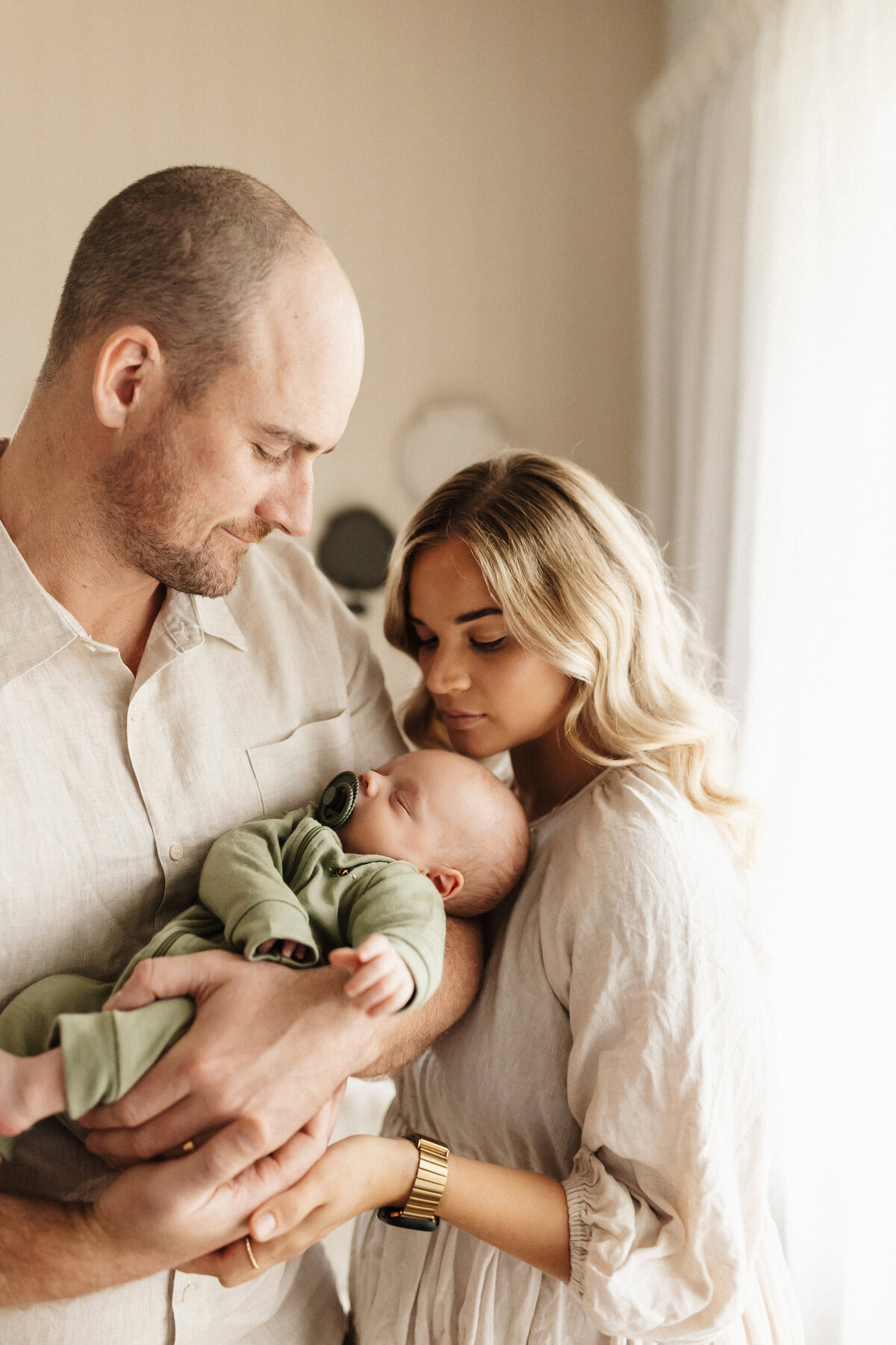 A mother and father holding their newborn baby who is wearing green while standing next to their window in their living room during their in-home newborn photoshoot with lucy jane photography in christchurch