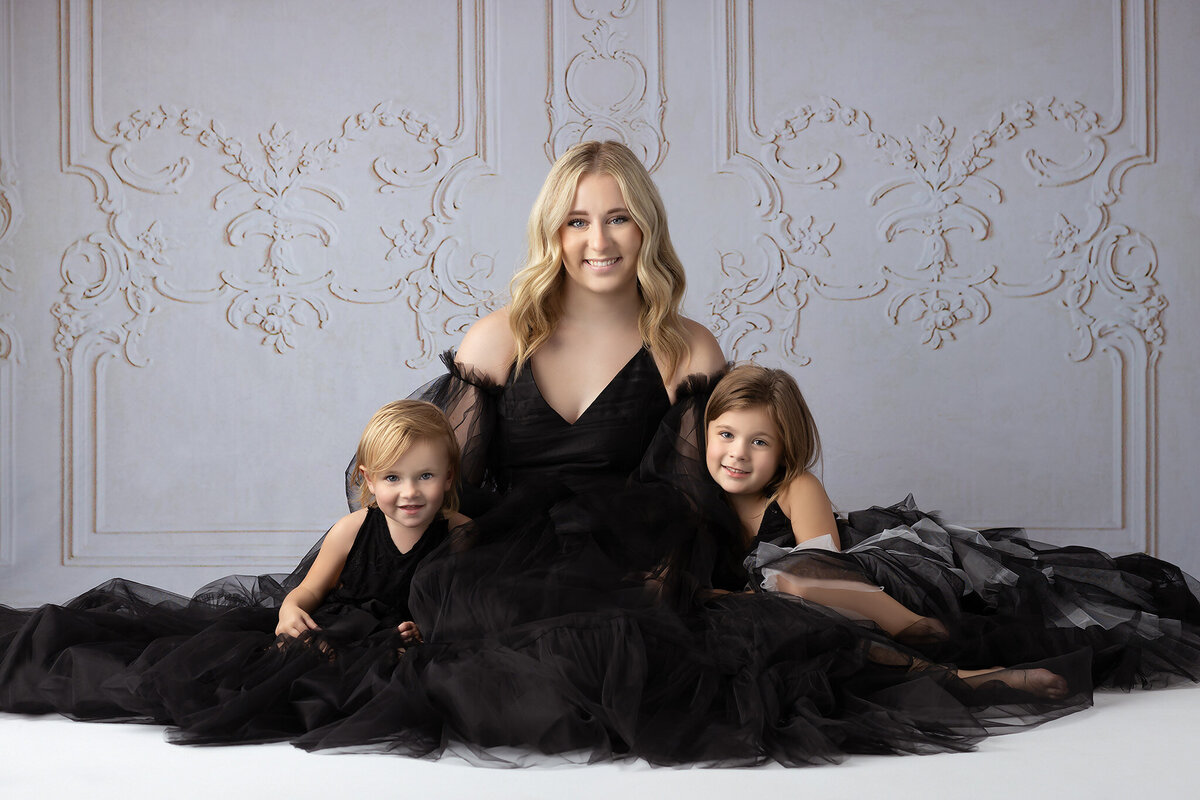 Sisters in black tulle dresses, seated together with soft smiles, posed in front of a classic backdrop.