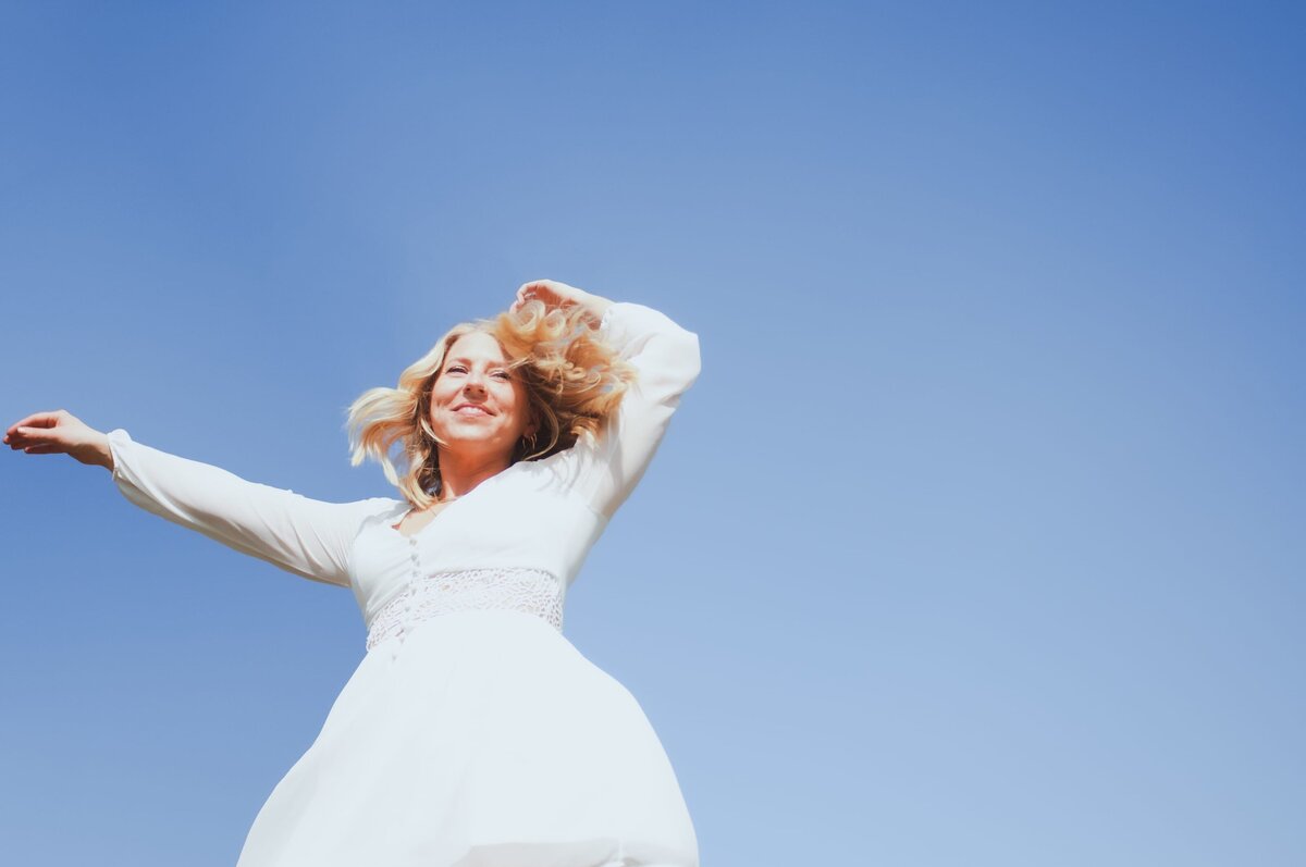 Blonde dances in white dress against blue sky background.
