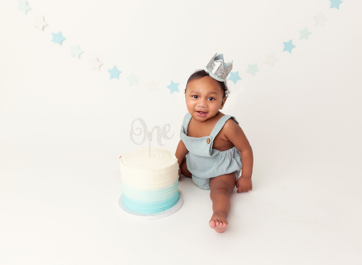 Baby boy poses with untouched cake before cake smash pictures in Brooklyn, NY. Baby is smiling wearing a blue overall romper and silver crown. Untouched blue ombre cake is between his legs.