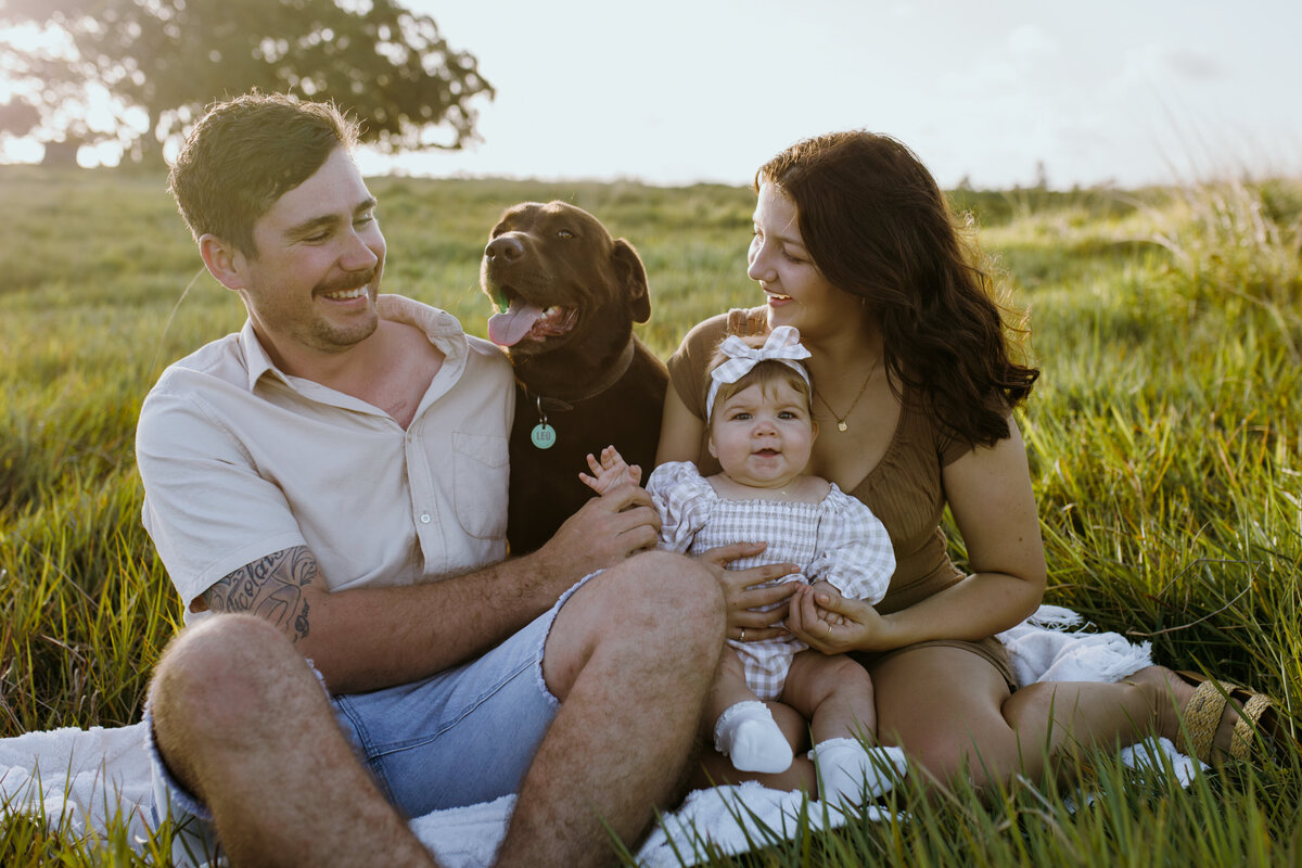 Family photo of one young child and their brown Labrador dog sitting in a sunlit grassy field.