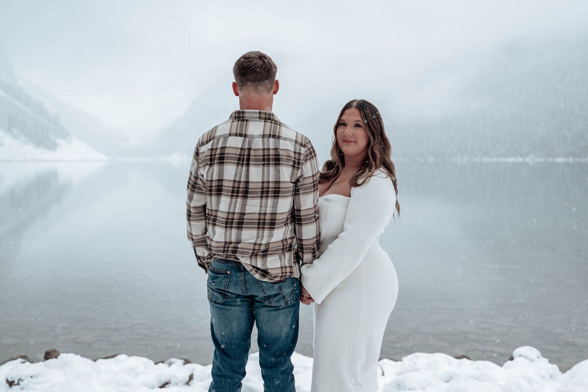 Bride-to-be smiles as fiancé looks out over the winter lake scene in Banff.