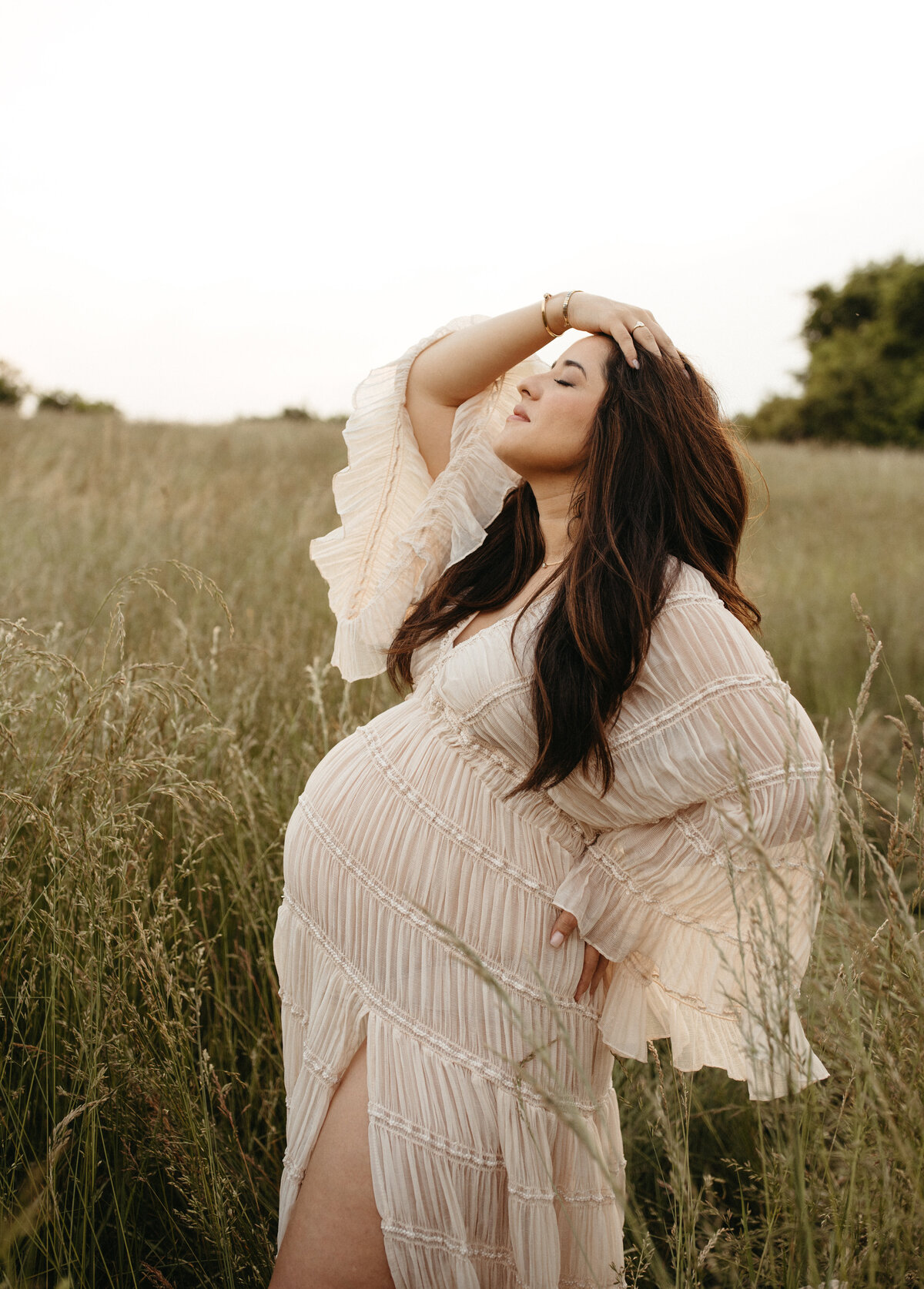 Pregnant woman wearing a flowy gown standing in a tall grass field during a golden-hour maternity session