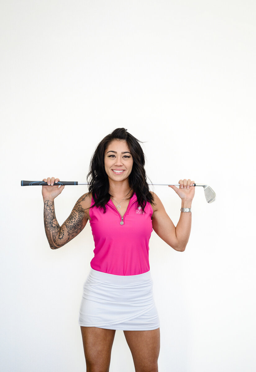 A golfer in a pink shirt holds a golf club over her shoulders and poses for branding photos and a branding picture session taken by a Denver branding photographer and brand photographer in Denver.