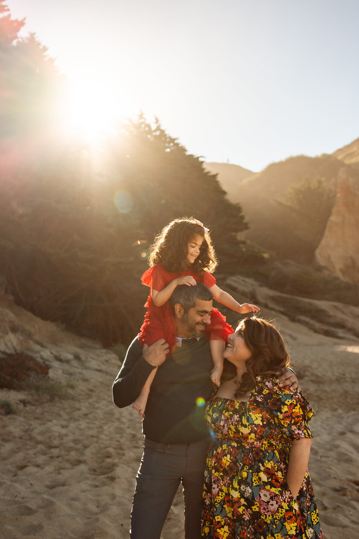 Family portrait on beach with girl on dad's shoulders and reaching for mom's head.  Warm sunlight in the background