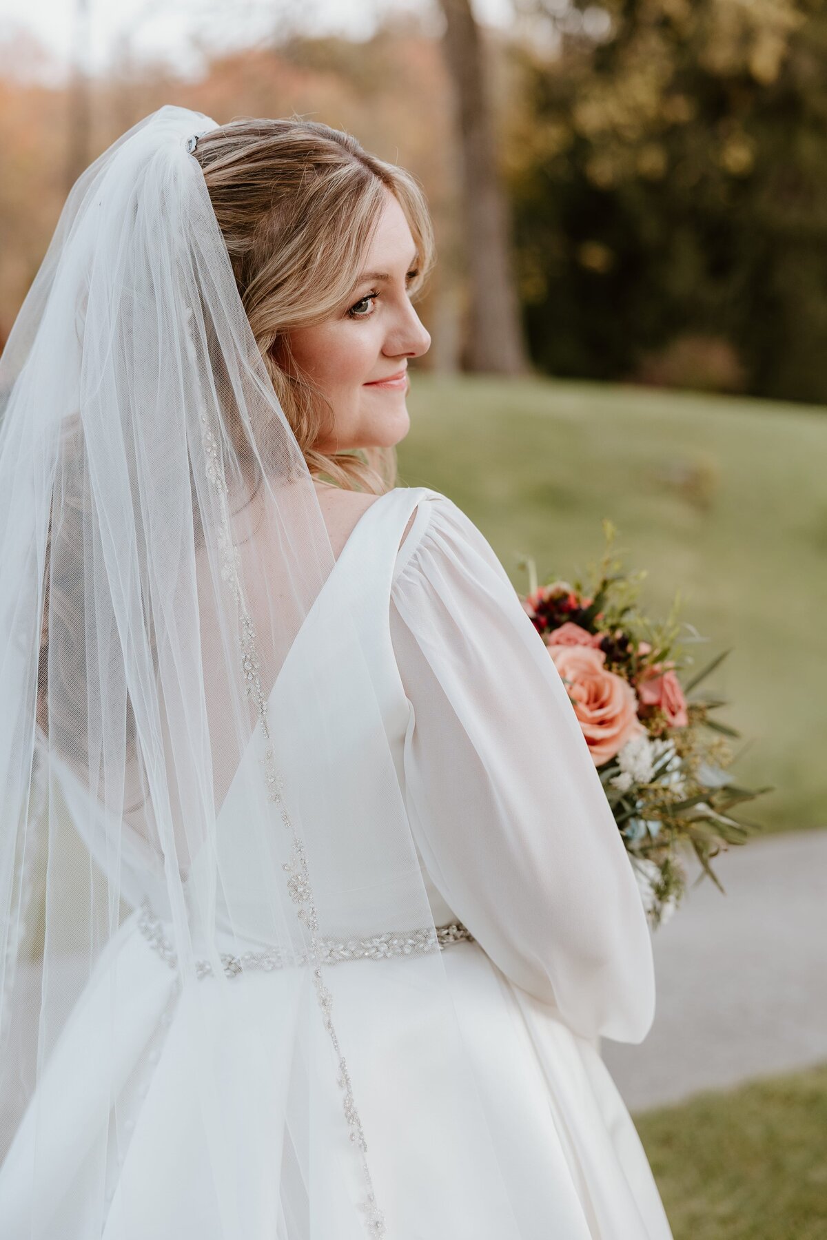 Bride softly smiling over her shoulder during golden hour, holding a bouquet of peach roses and greenery, wearing a long veil and flowy-sleeved wedding gown with delicate beading on the back, photographed outdoors in the Hudson Valley.