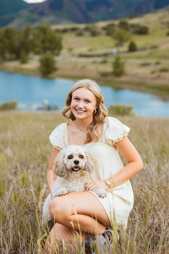 Senior girl squats with dog in her lap in front of a blue lake.
