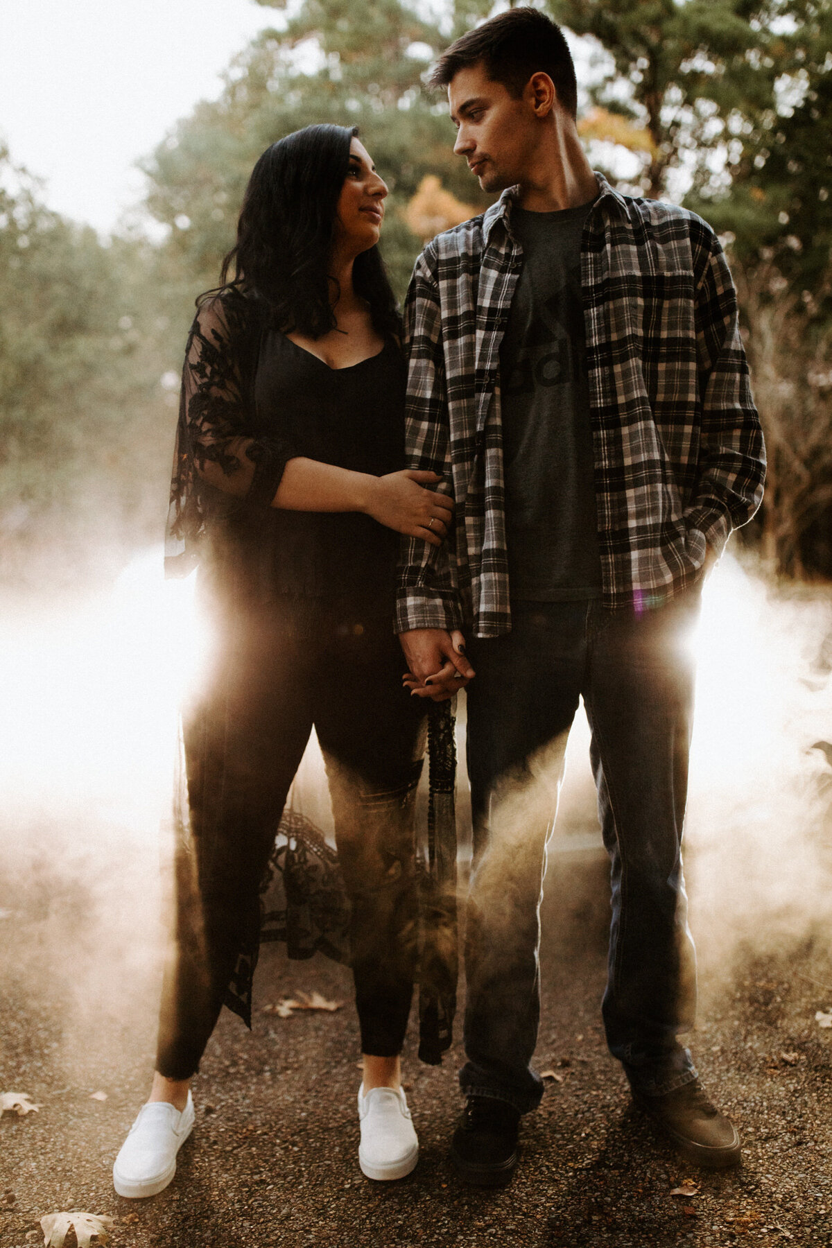Guy wearing black Vans and a grey flannel holding hands with a girl wearing all black while they look at each other and stand in front of a car with headlights shining through the fog