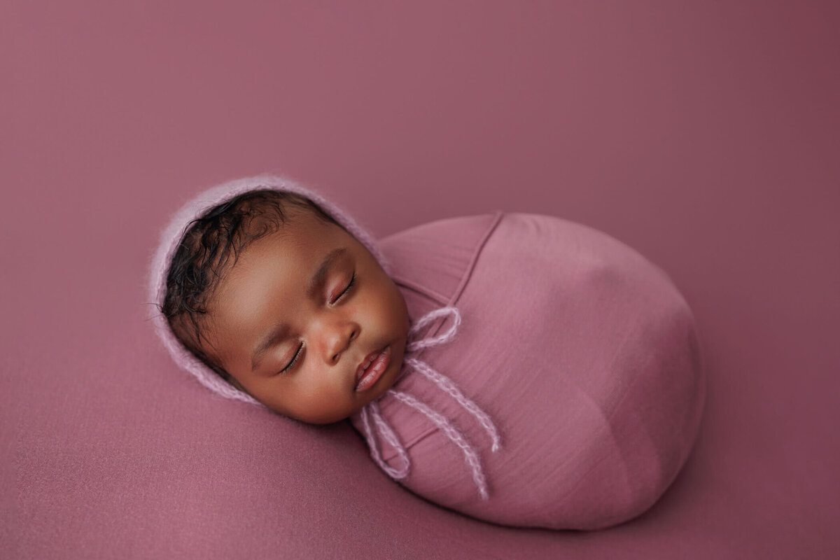 Sleeping newborn baby girl wrapped in a soft pink blanket with a matching pink bonnet, lying on a pink backdrop.