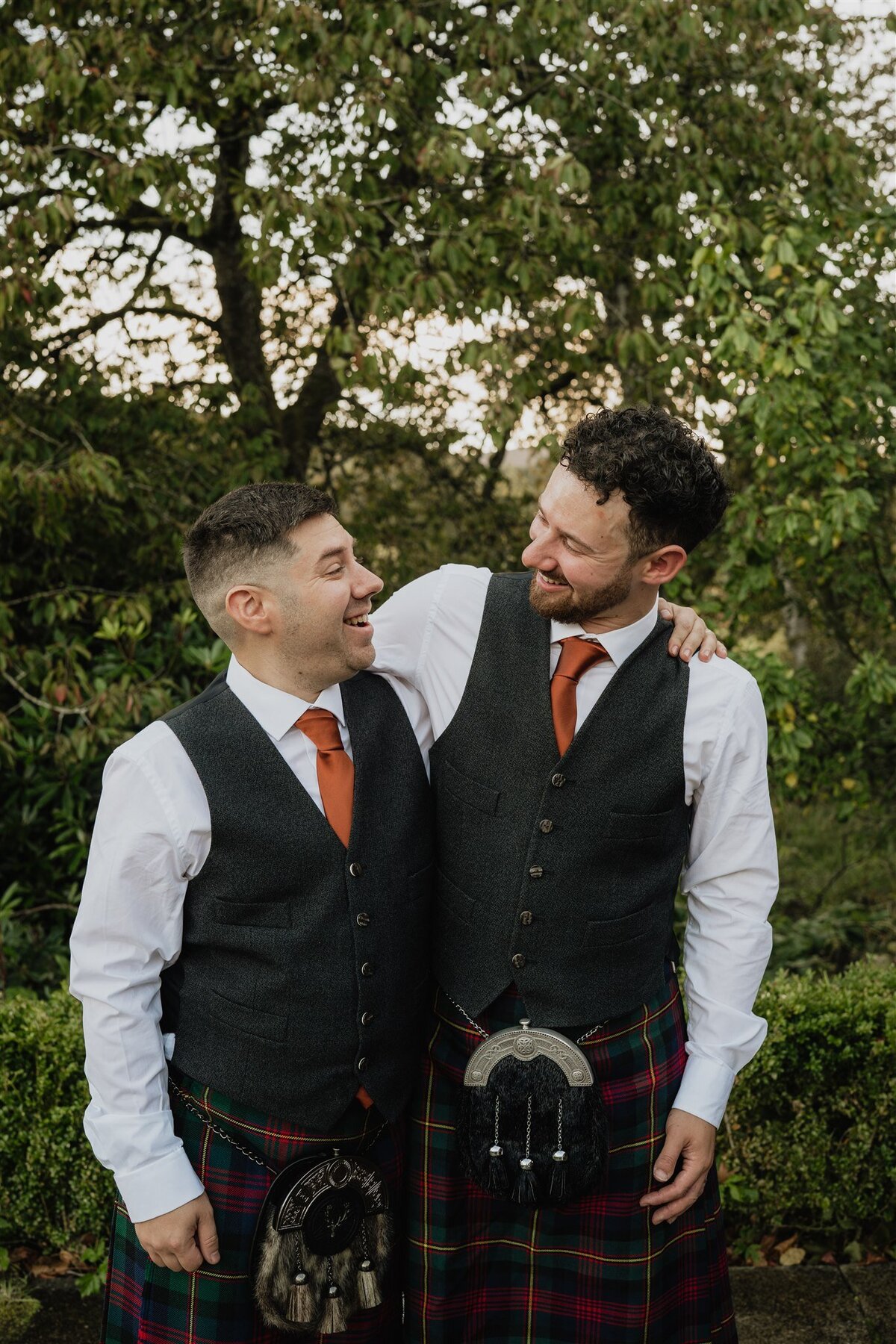 Two men stand side by side and pose for a photograph. They both wear kilts and traditional Scottish wedding attire. Trees and greenery surround them.