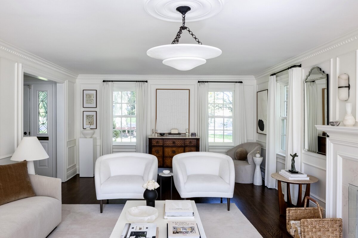 Elegant and sophisticated living room designed by Deni + Dove Interiors, a New Jersey-based interior design studio serving Short Hills, Chatham, Montclair, Summit, Ridgewood, and surrounding areas. This space features a neutral color palette with white upholstered chairs, a beige sofa with a brown accent pillow, a central coffee table styled with books and vases, and a vintage wooden console. Tall windows with flowing white curtains, decorative wall paneling, and a fireplace with sculptural decor create a timeless and inviting ambiance.