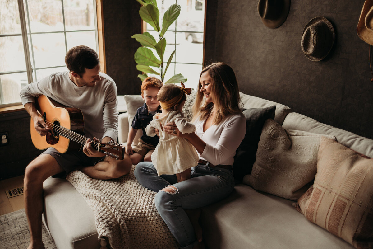 Family of four hang out in well designed living room while dad plays the guitar and mom helps toddler daughter dance