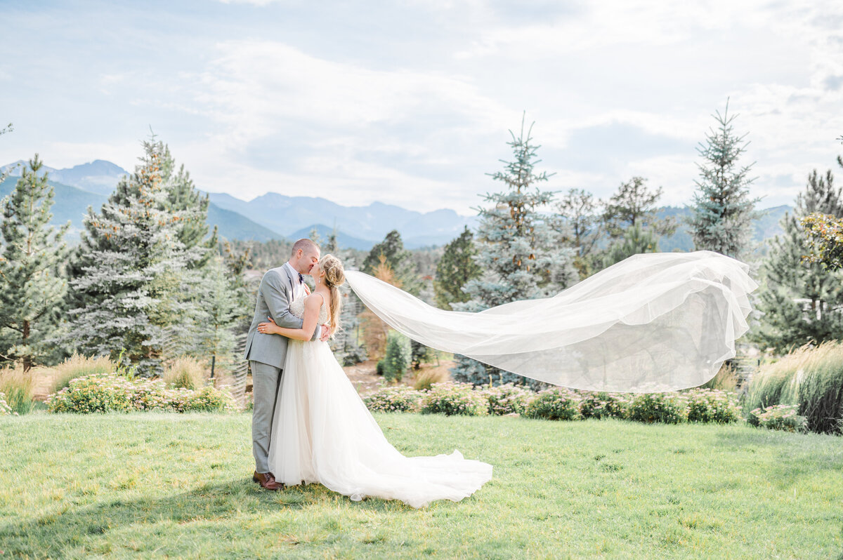couple kisses as the brides veil flies behind her in the wind on the grassy lawn at the stanley hotel in estes park colorado