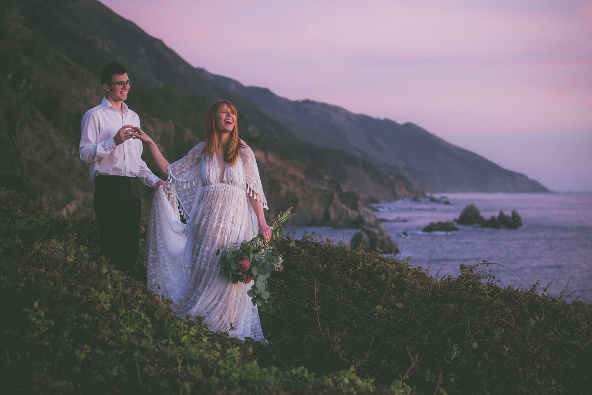 Bride and groom hold hands with beautiful Big Sur scenery in background.