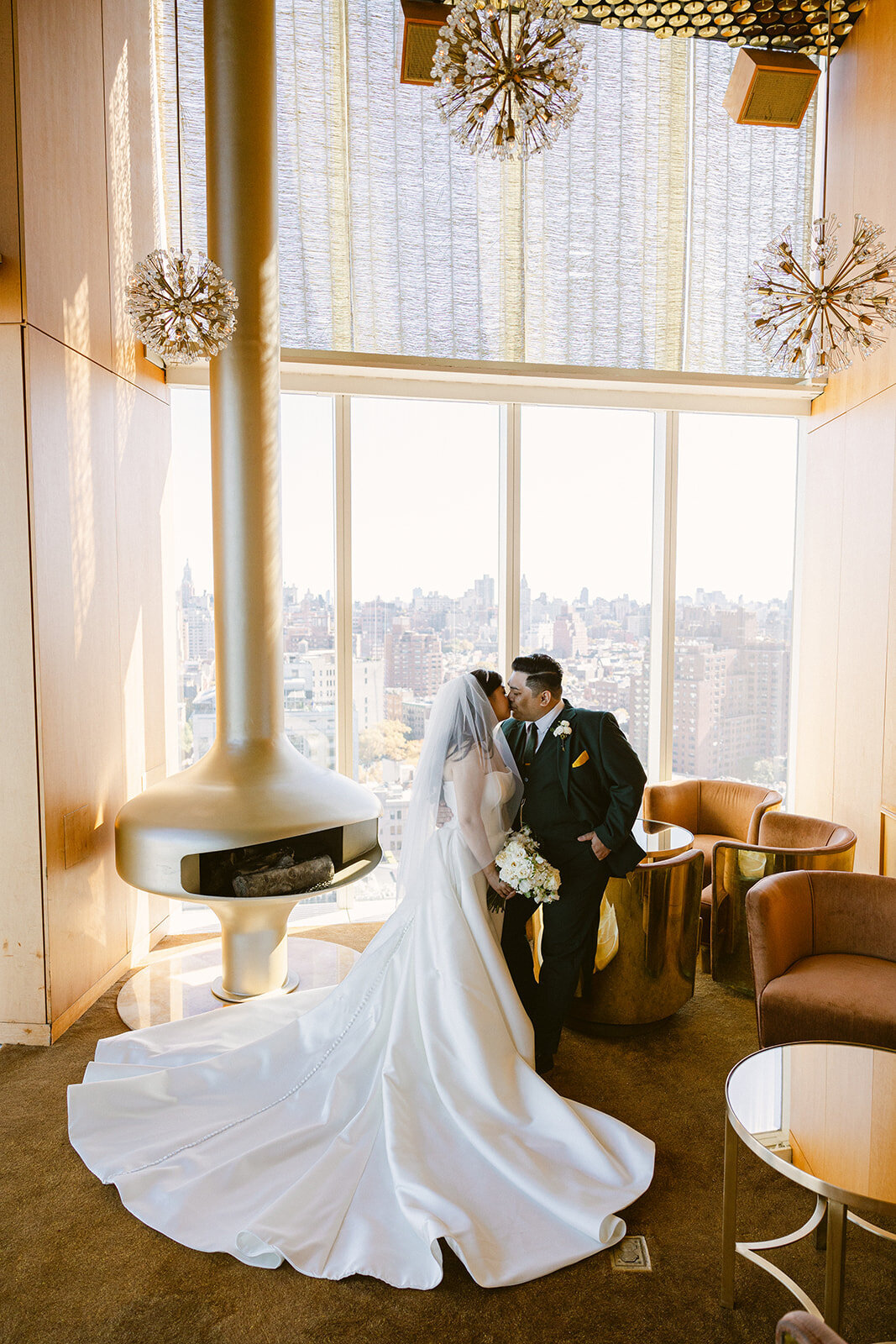 A bride and groom share an intimate moment in a modern, luxurious lounge with floor-to-ceiling windows overlooking a city skyline.