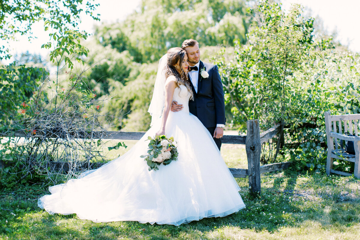 Bride and groom embracing with eyes closed, standing by a rustic old fence as golden sunlight soaks them in. Captured at Minnesota Landscape Arboretum by Toly Dzyuba Photography.