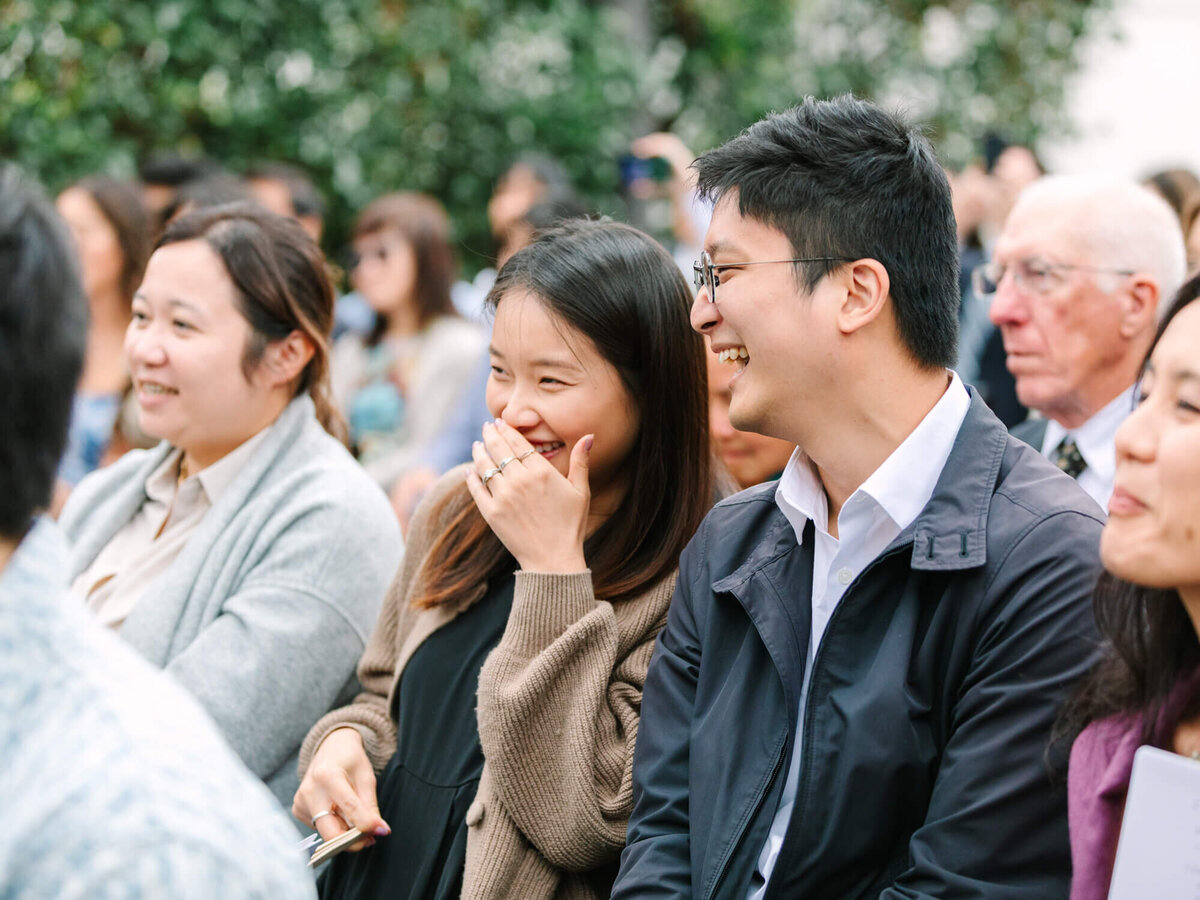 Guests sitting outdoors, smiling and laughing during a wedding ceremony at Muckenthaler Mansion in Orange County. They appear engaged and happy, with blurred greenery in the background. Casual and cheerful atmosphere.