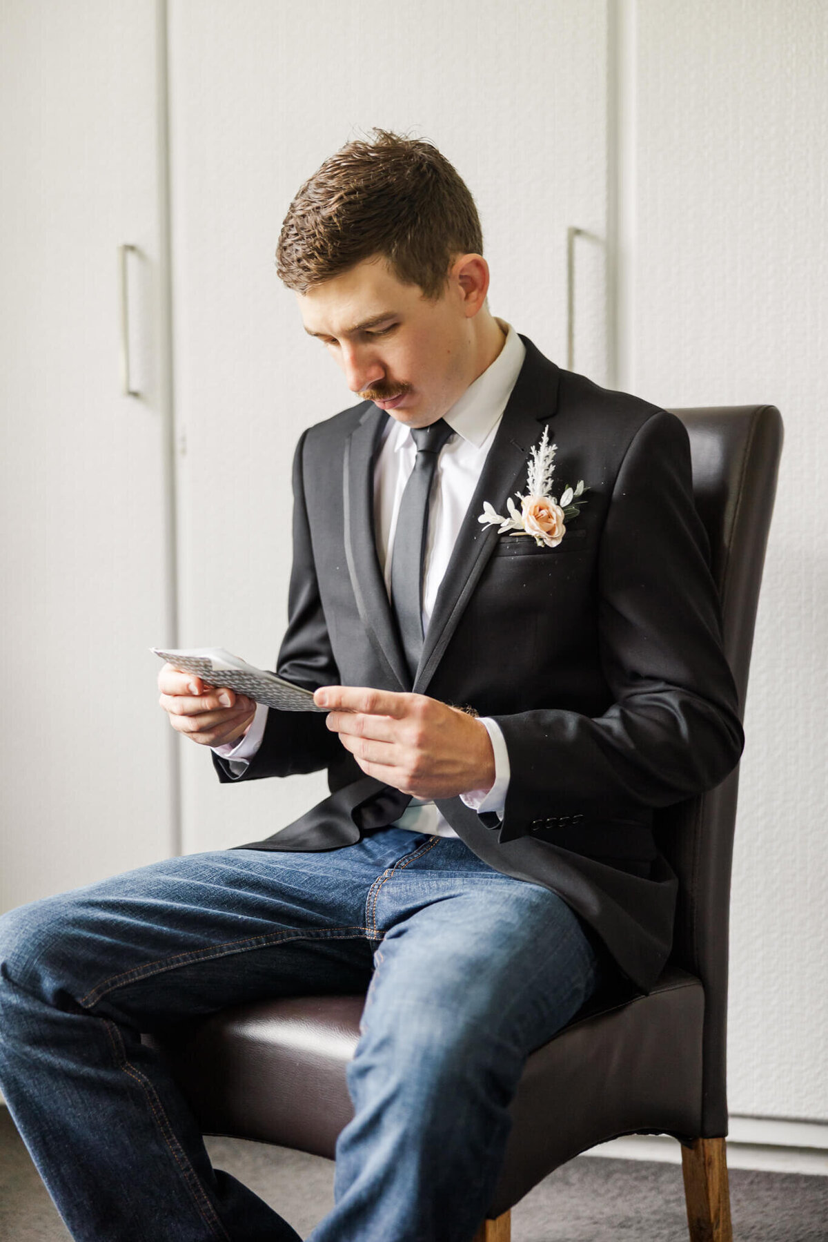 Groom sits on a dark brown seat and reads a note from his bride before the wedding ceremony.