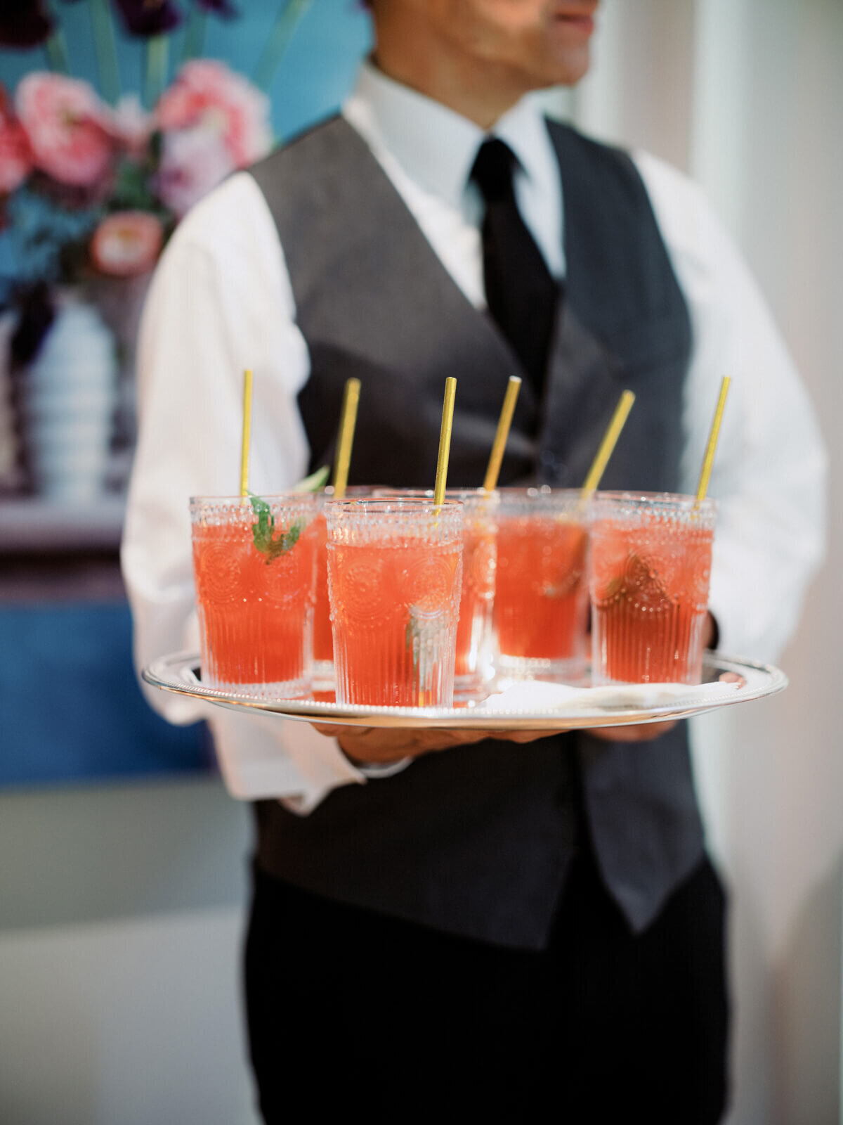 The waiter is holding a round tray of six red drinks with straws.
