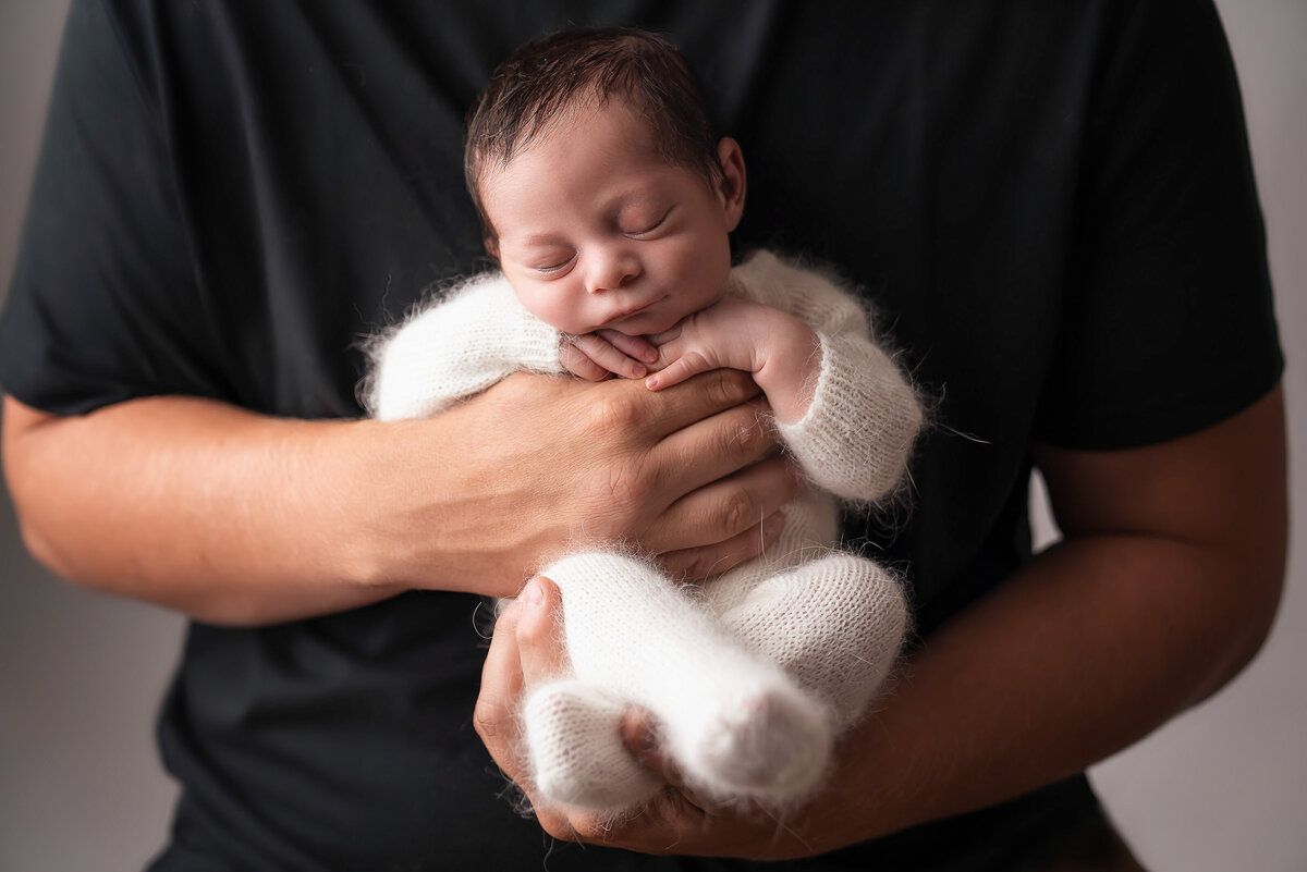 Dad is holding his sleeping baby boy against his chest. Only dad's arms are visible. Baby's hands are folded atop of Dad's fingers. Captured by Bryn Mwar Philadelphia and Main Line photographer Liza Nicole.