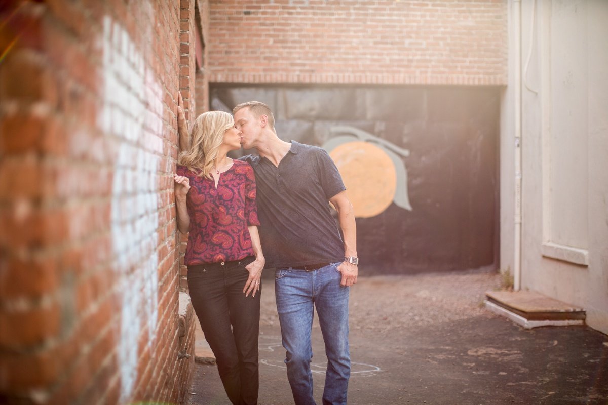 Future Bride and Groom share a kiss during an engagement session in a back alley as she leans against an old brick wall