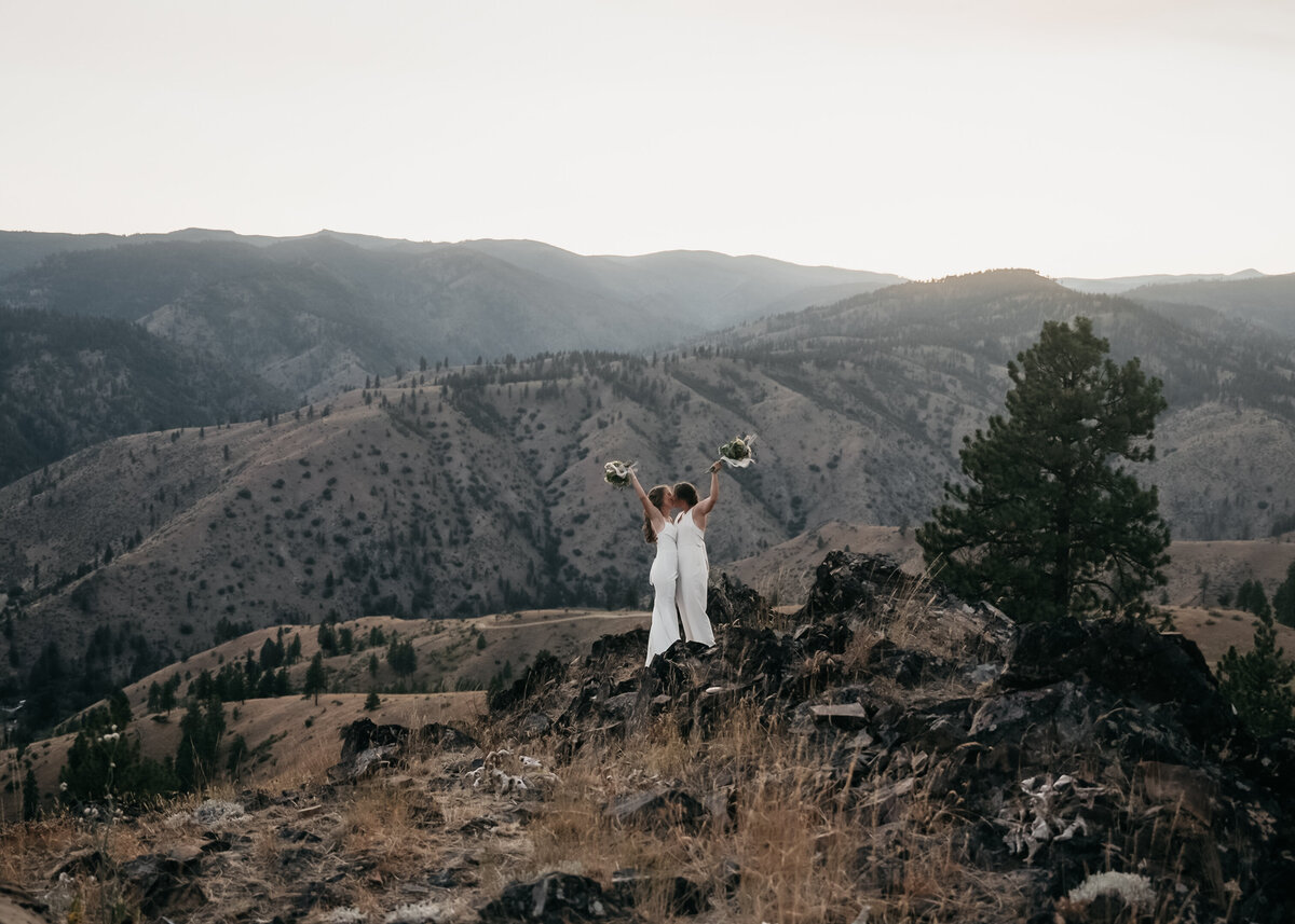 Two brides, dressed in elegant wedding gowns, share a kiss inside a fire lookout with breathtaking mountain views.