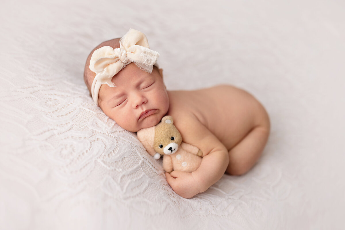 Newborn girl lying on her side on a white textured blanket wearing a cream bow headband.
