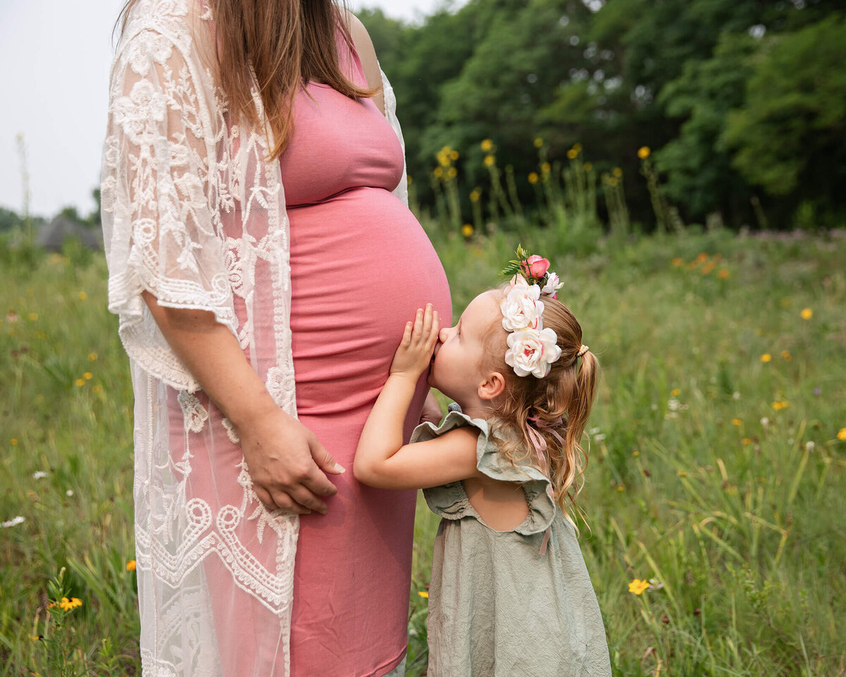 Girl kissing her Mom's baby bump in a field of flowers.