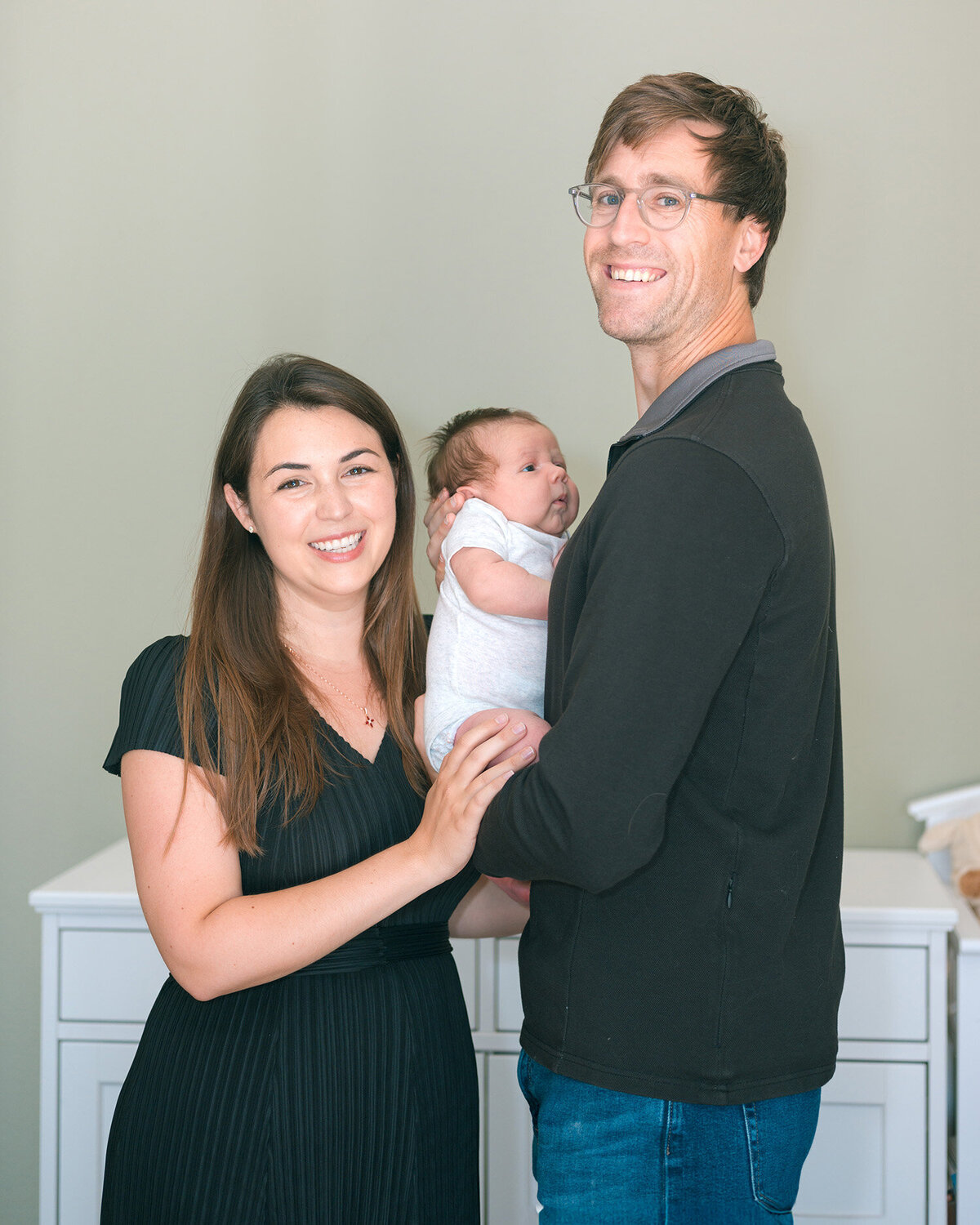 Young parents wearing black smiling while holding newborn baby in white onesie during in-home session, captured by Menlo Park newborn photographer.
