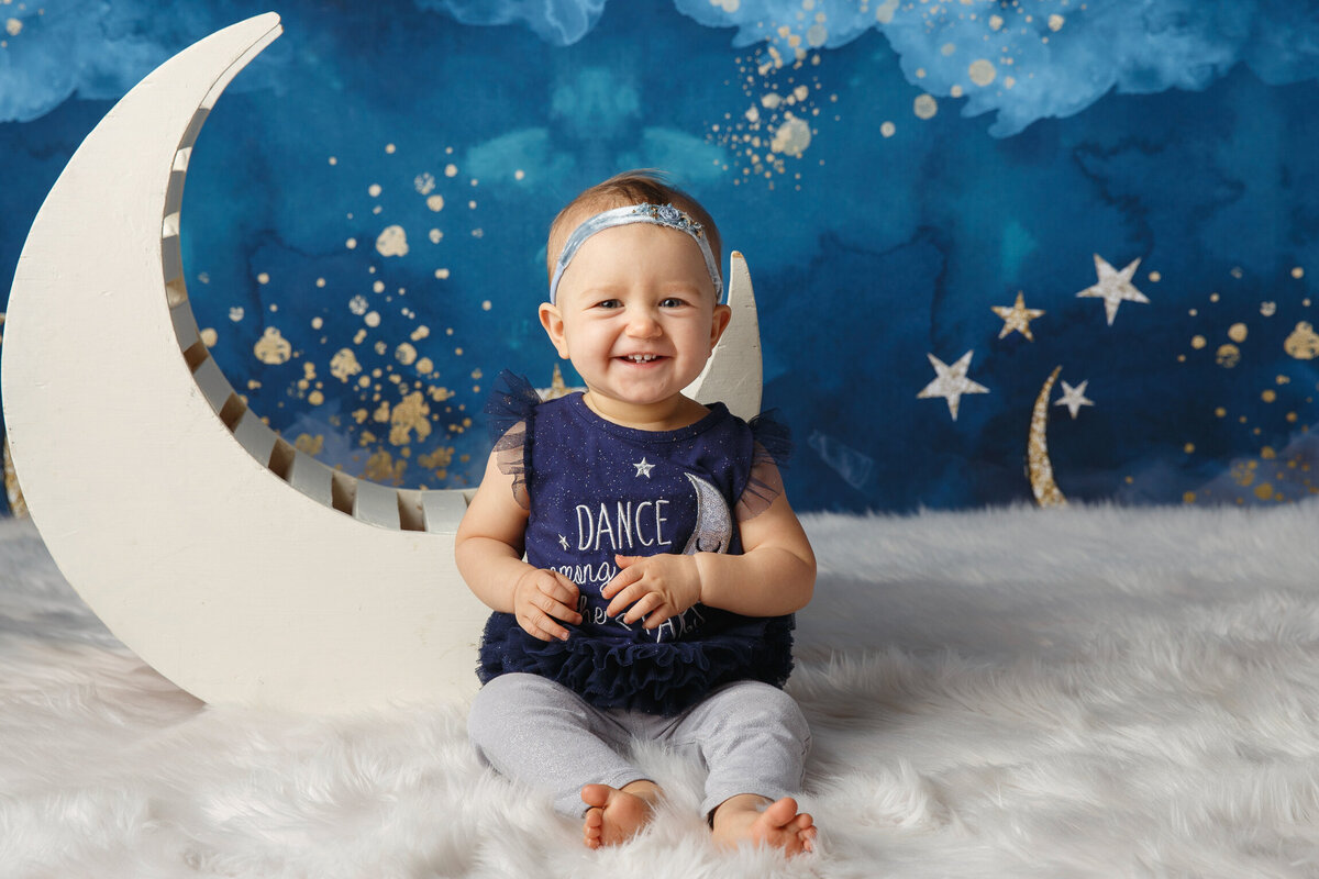 Cute baby girl sitting next to a wooden moon prop with a starry sky background
