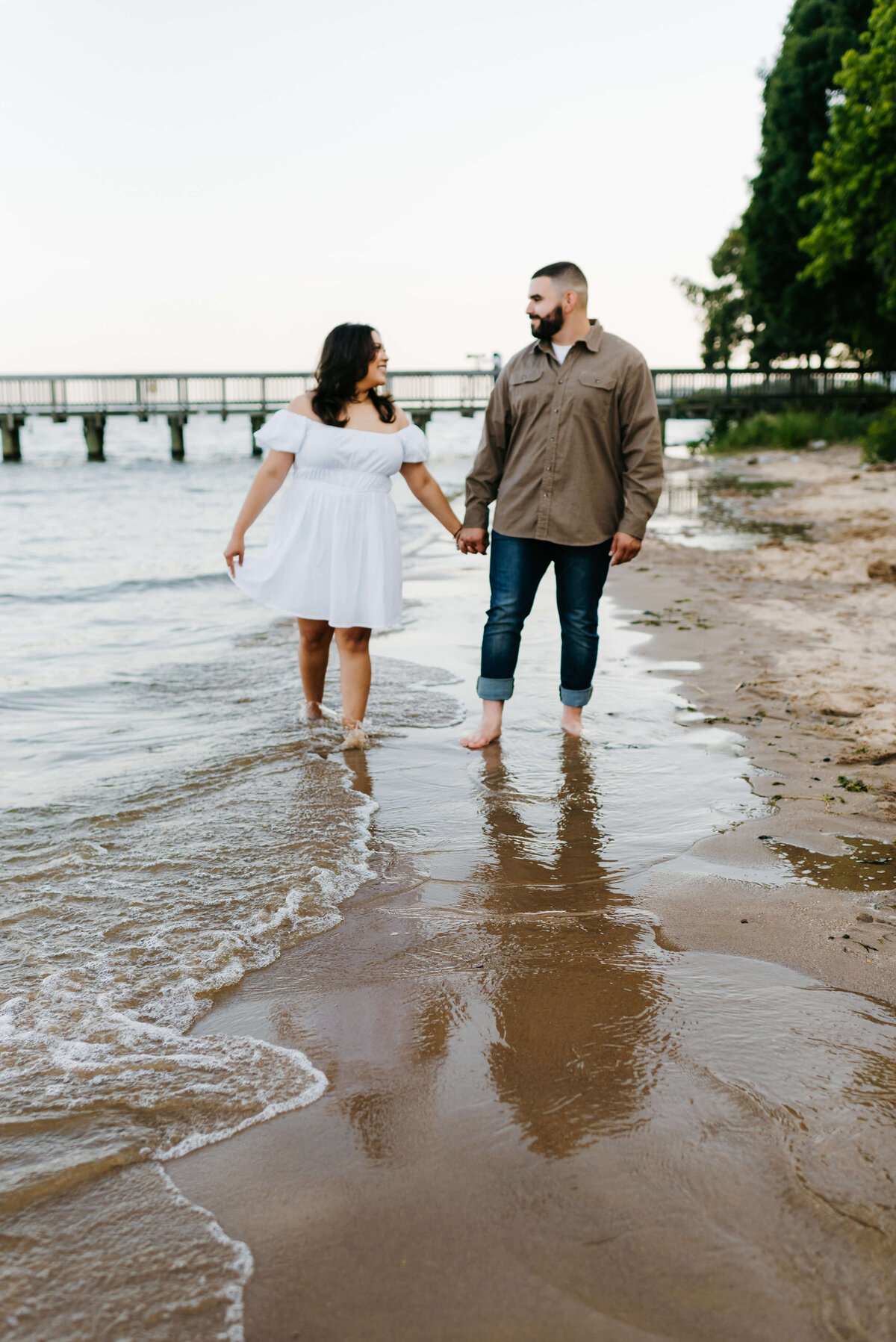 couple holding hands as they walk in the shallow water at a beach  and smile at one another for couples photography