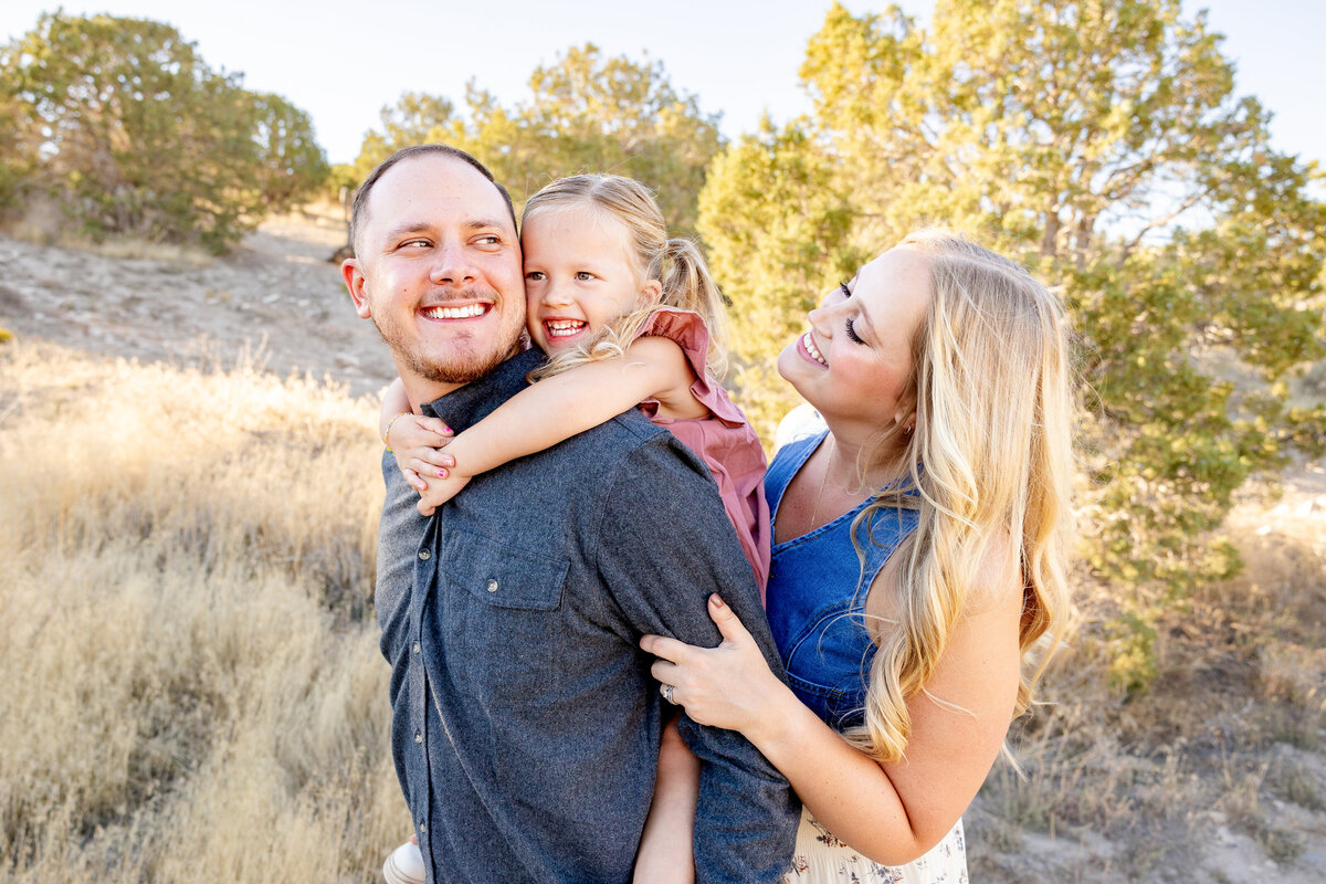 A father carries his daughter on his back while the mother walks beside them, all smiling joyfully.  Taken by Cassie Lee Photography