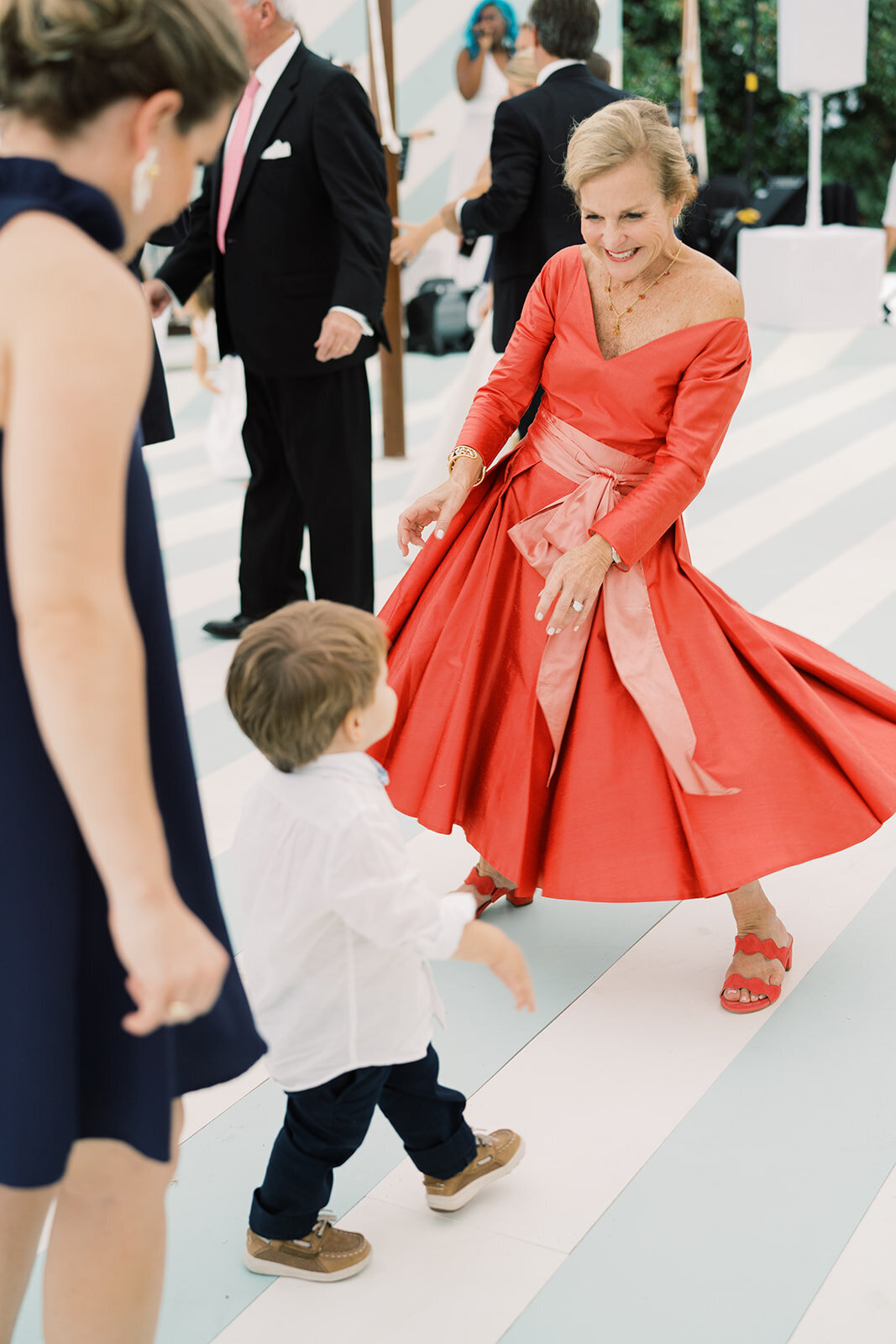 The joyful moment captured in the photo was taken at The Hamptons, New York, waterfront estate tented wedding by photographer Bonnie Sen, with videographers East West Films also capturing the beautiful moments. The image shows a happy elderly woman in a red dress twirling on a striped dance floor, hand in hand with a young boy in a white shirt and dark pants. In the background, other adults dressed in formal attire can be seen, adding to the festive atmosphere of the celebration.