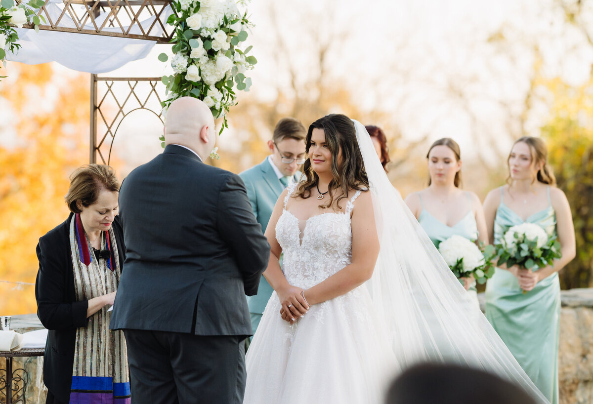outdoor wedding ceremony with bride holding her hands together while standing under an arch with her bridesmaids in blue bridesmaids dresses standing behind her as she smiles at her groom captured by Virginia wedding photographer