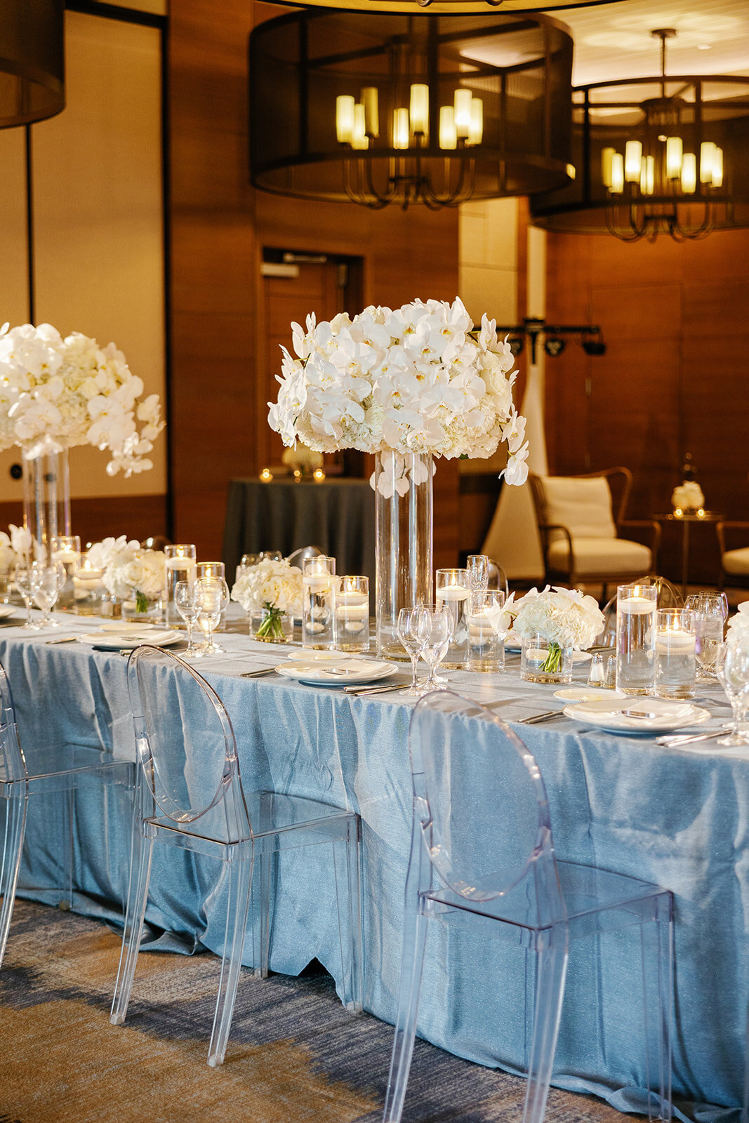 A beautifully set wedding reception table featuring tall white floral centerpieces, clear chairs, and elegant lighting at a Miami wedding venue, captured by Claudia Amalia Photography.