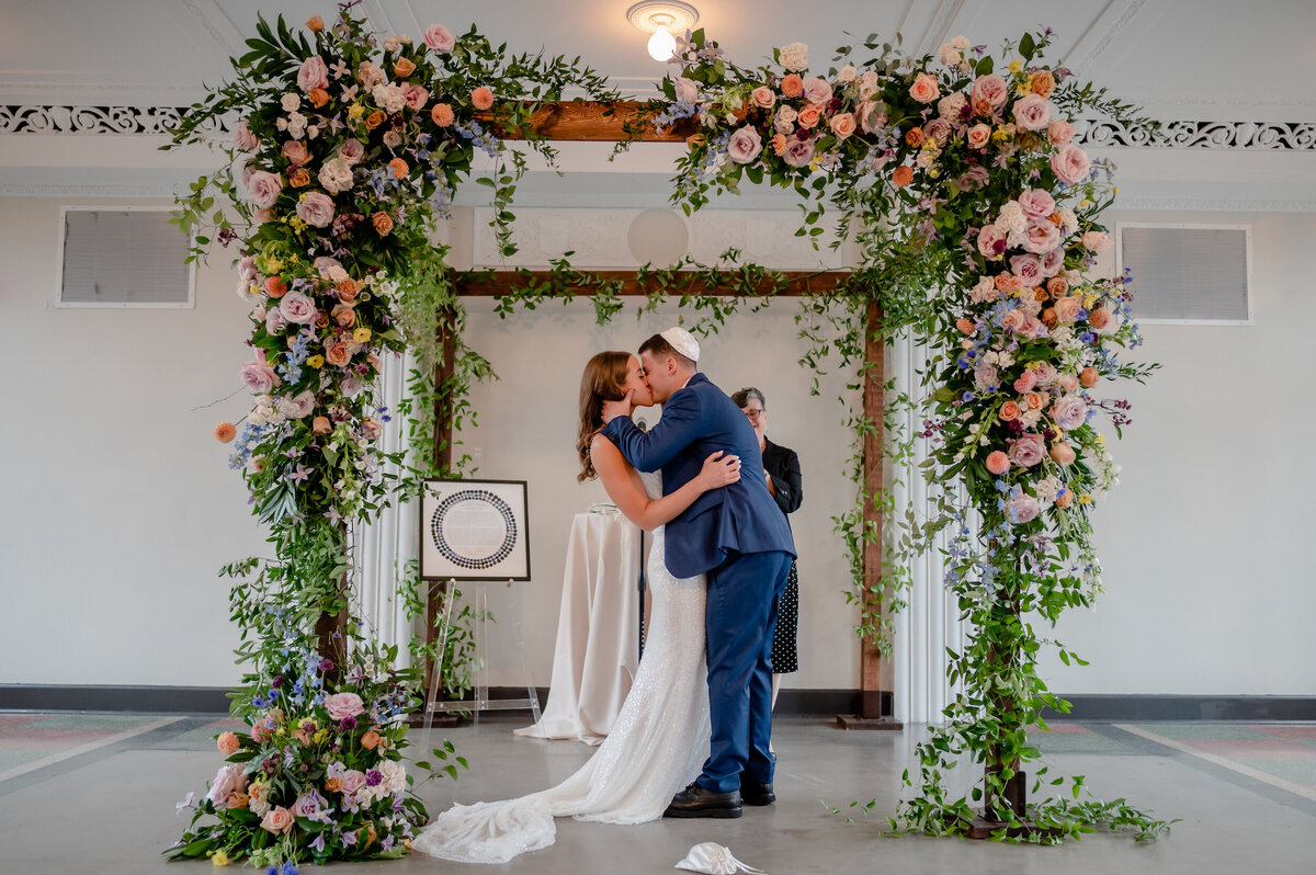 Pink flowers cover the chuppah while couple share their first kiss at Revel Motor Row wedding