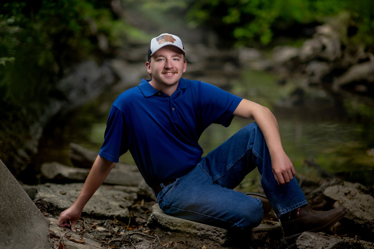 A high school senior in jeans and blue polo and hat sits on rocks in a forest smiling for his Pella Senior Photographer