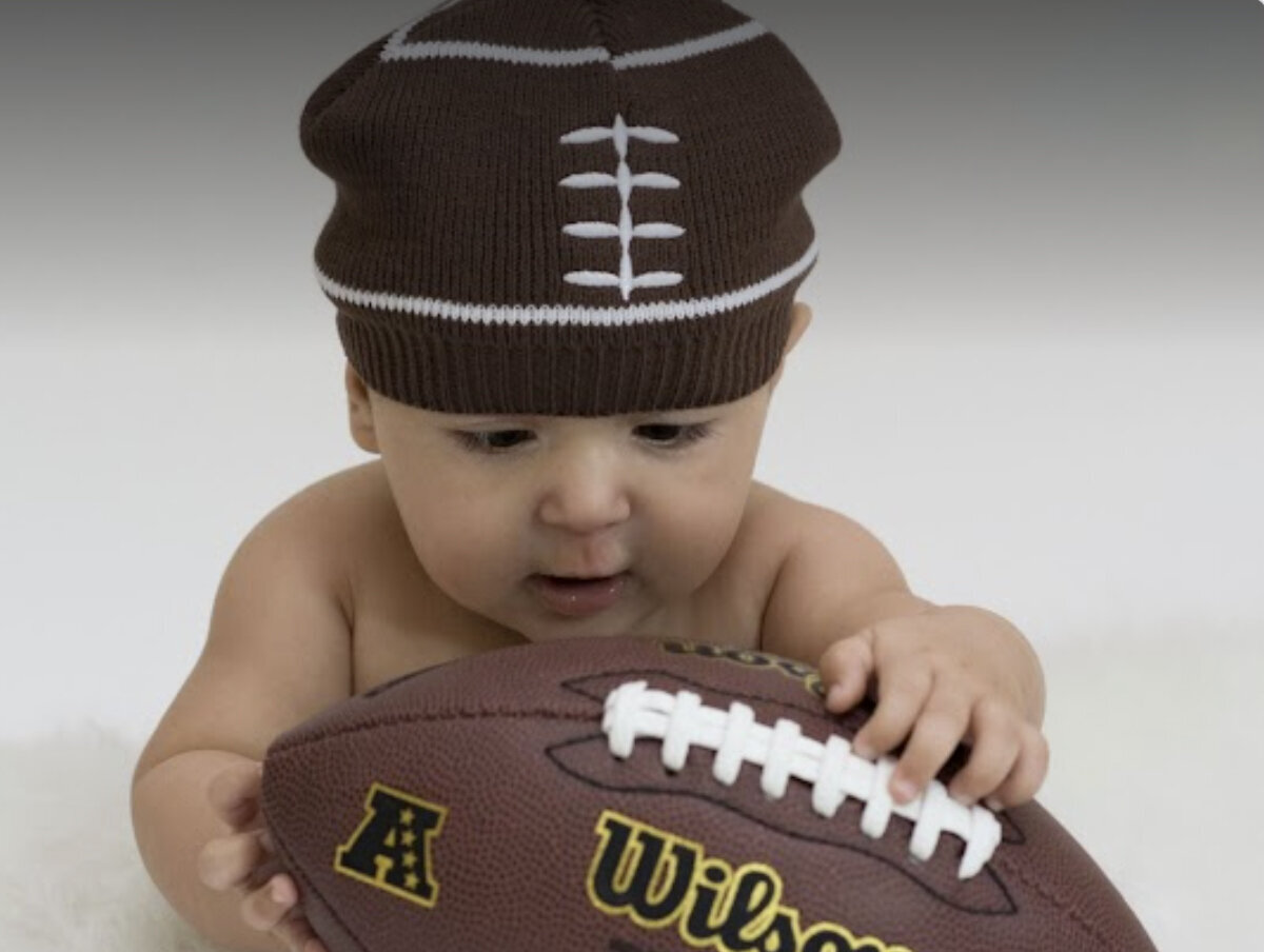 An image from an Orange County milestone photography studio of a baby playing with a football wearing a football beanie