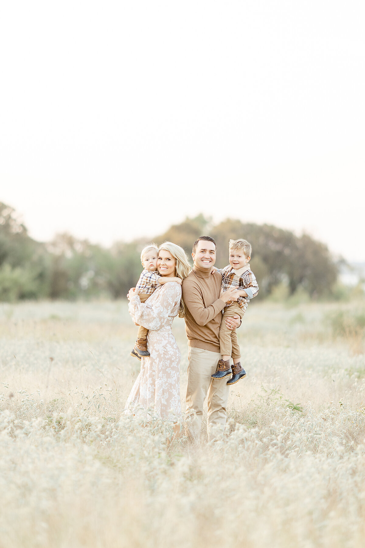 Beautiful family portrait of a mother and father holding their two young boys as they look and pose for family photos in the middle of an open field at a DFW park.