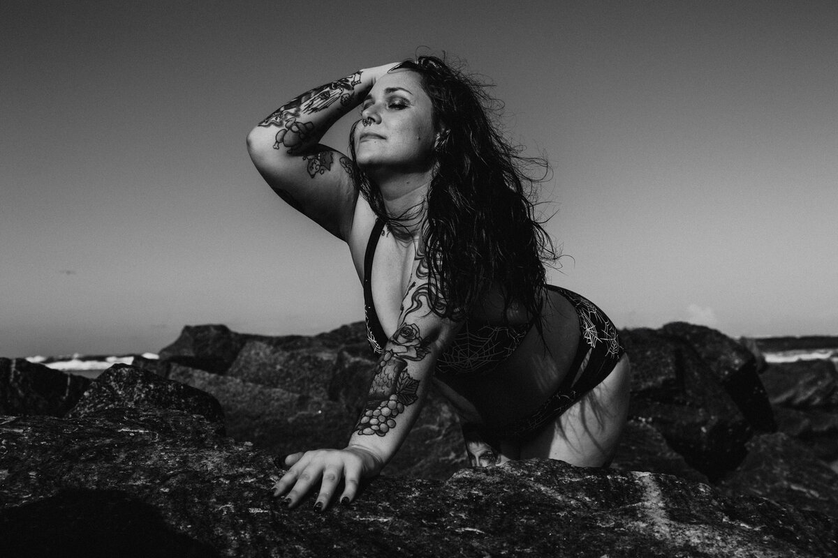 Tattooed woman posing confidently on ocean rocks in a spiderweb bikini during a Salted Summer beach boudoir shoot in Jacksonville.