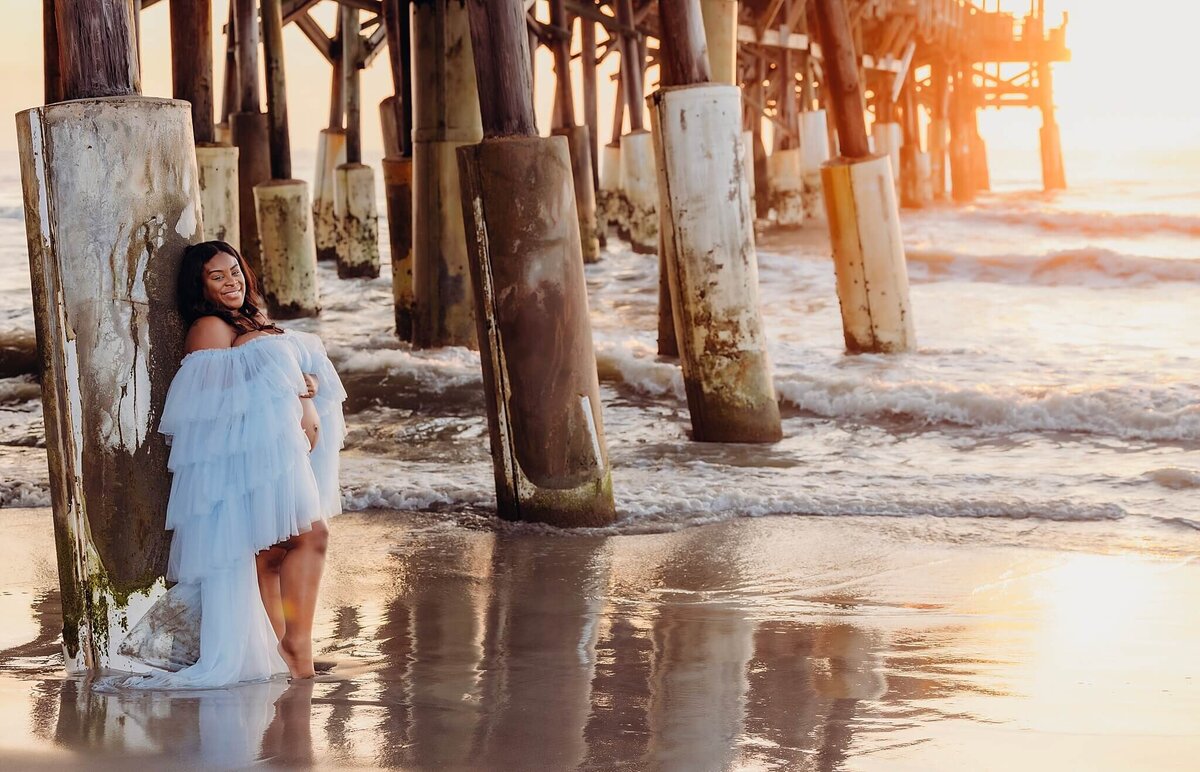 A pregnant woman in a flowing blue dress with ruffles stands with her back against a pier piling. The pier is located on a sandy beach with foamy waves lapping at the shore. The setting sun casts a warm glow on the scene. The woman is smiling and appears to be enjoying the warm evening light. The setting creates a peaceful and serene atmosphere, perfect for a maternity photoshoot.