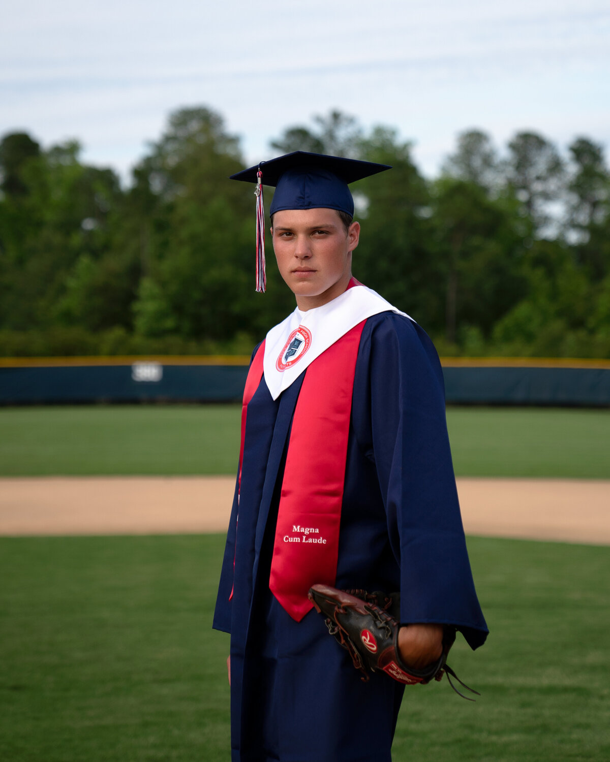 Classic serious face portrait with Connor Resavy in cap and gown with baseball glove on left hand. Captured at Apex-Friendship High School in Spring 2020