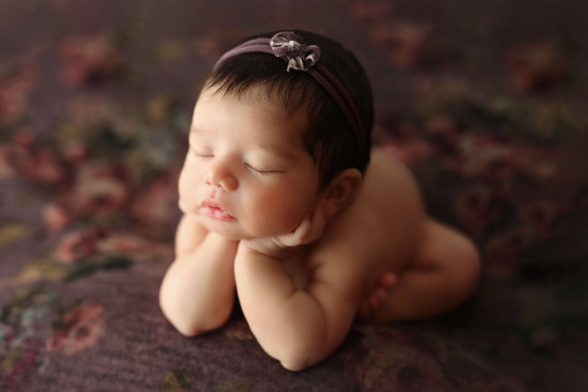 baby girl in froggy pose with a purple headband on a floral purple backdrop at her newborn photography session with a photographer at a reston va photo studio