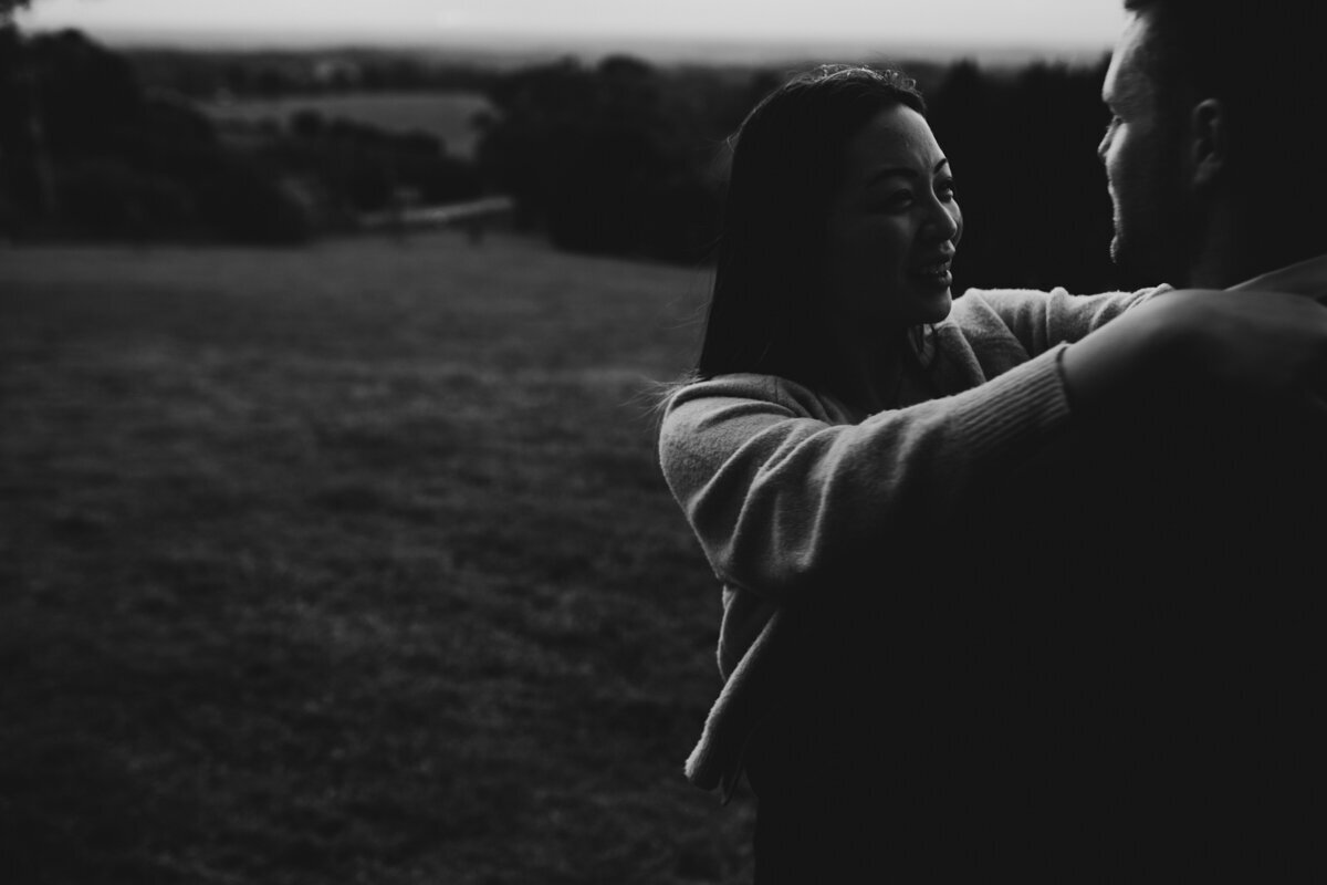 Bride and Groom standing on a mountain top. The wind is blowing in her hair and they are looking out to the view while hugging each other. The bride is a long sleeved wedding gown.